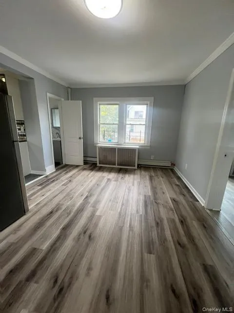 Unfurnished bedroom featuring ornamental molding, freestanding refrigerator, wood finished floors, radiator, and a baseboard heating unit Unfurnished bedroom featuring ornamental molding, freestanding refrigerator, wood finished floors, radiator, and a baseboard heating unit