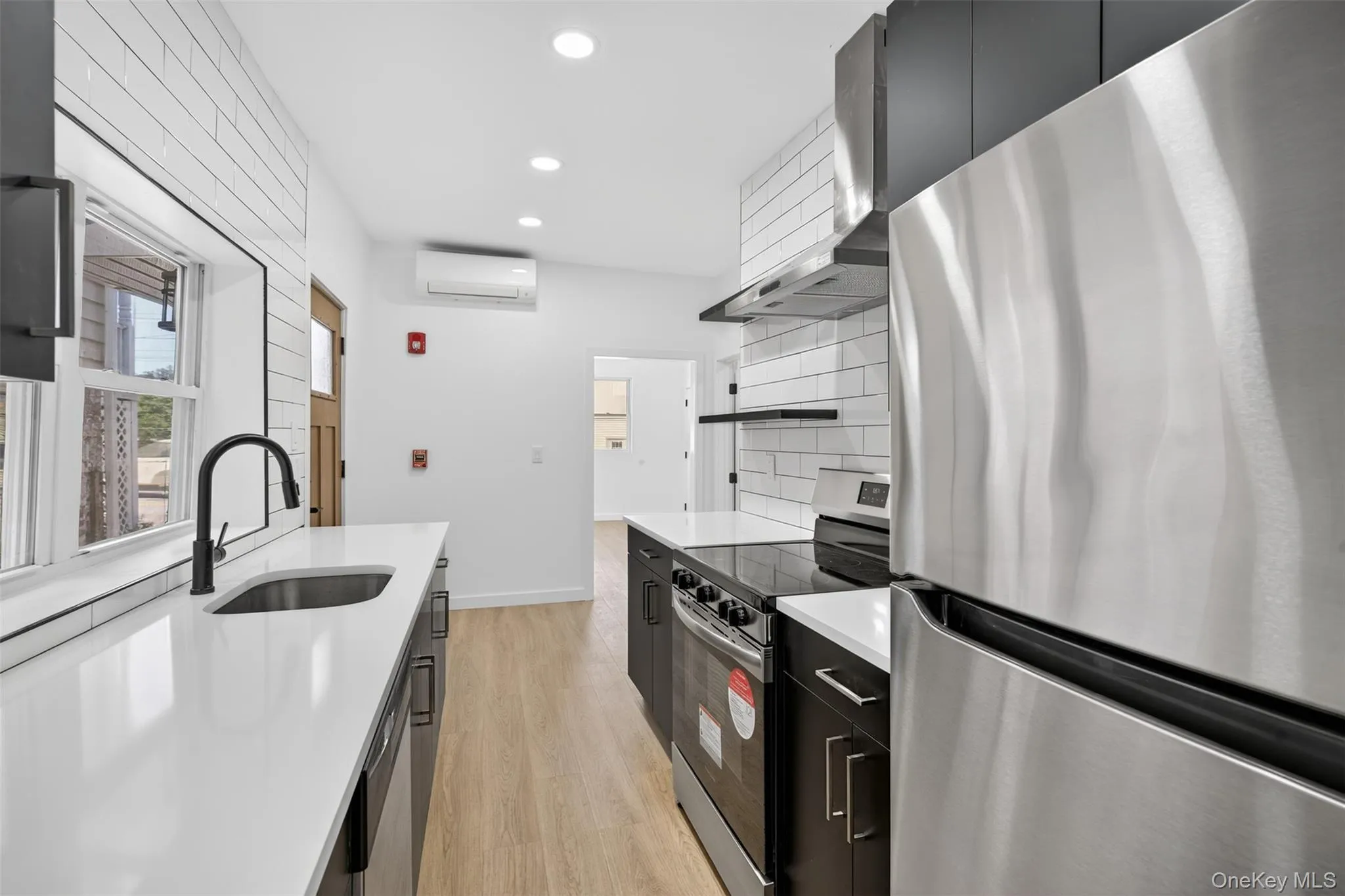 Kitchen featuring dark cabinetry, stainless steel appliances, light wood-type flooring, wall chimney exhaust hood, and light stone countertops Kitchen featuring dark cabinetry, stainless steel appliances, light wood-type flooring, wall chimney exhaust hood, and light stone countertops