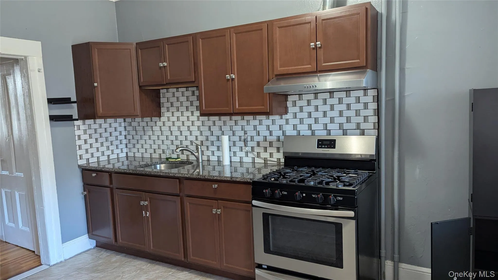 Kitchen with gas stove, tasteful backsplash, dark stone counters, under cabinet range hood, and brown cabinetry Kitchen with gas stove, tasteful backsplash, dark stone counters, under cabinet range hood, and brown cabinetry