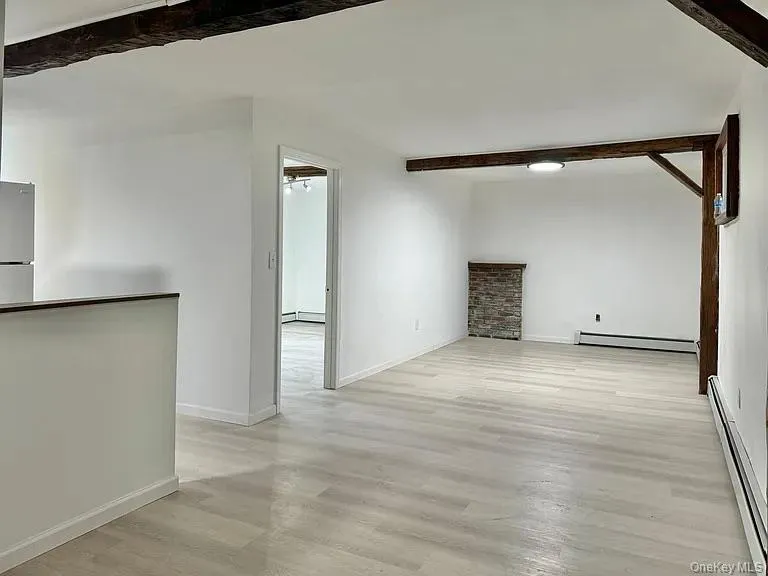 Unfurnished living room with beamed ceiling, a baseboard heating unit, light wood-type flooring, and a baseboard radiator Unfurnished living room with beamed ceiling, a baseboard heating unit, light wood-type flooring, and a baseboard radiator