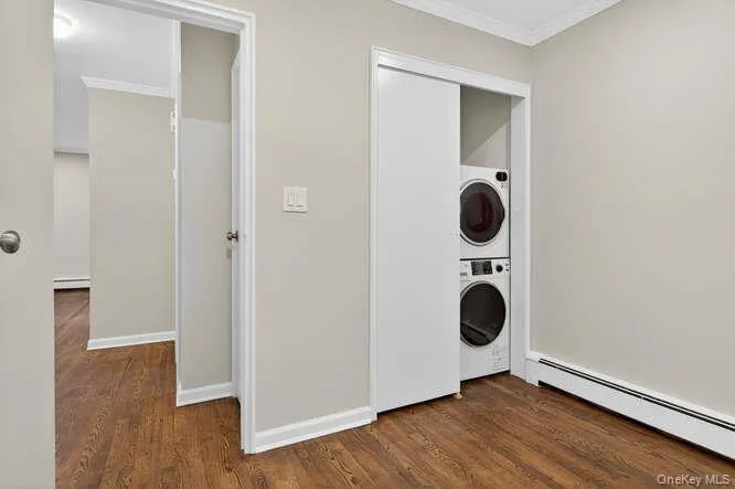 Laundry room with a baseboard heating unit, dark wood-type flooring, ornamental molding, and stacked washer / dryer Laundry room with a baseboard heating unit, dark wood-type flooring, ornamental molding, and stacked washer / dryer