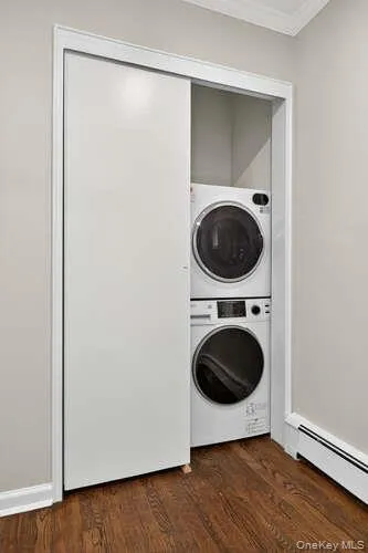 Washroom with baseboard heating, estacked washer and dryer, dark wood-style flooring, and crown molding Washroom with baseboard heating, estacked washer and dryer, dark wood-style flooring, and crown molding