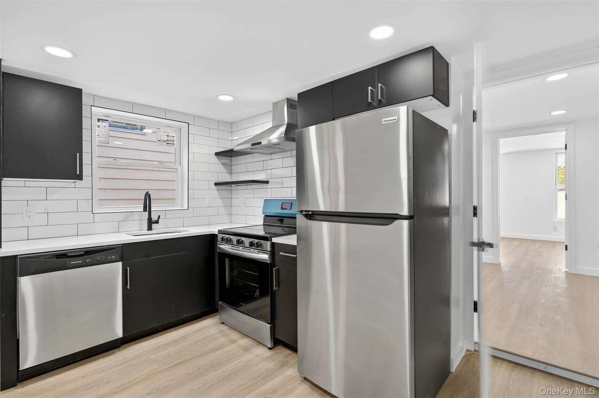 Kitchen with dark cabinets, appliances with stainless steel finishes, decorative backsplash, open shelves, and light wood-style floors Kitchen with dark cabinets, appliances with stainless steel finishes, decorative backsplash, open shelves, and light wood-style floors