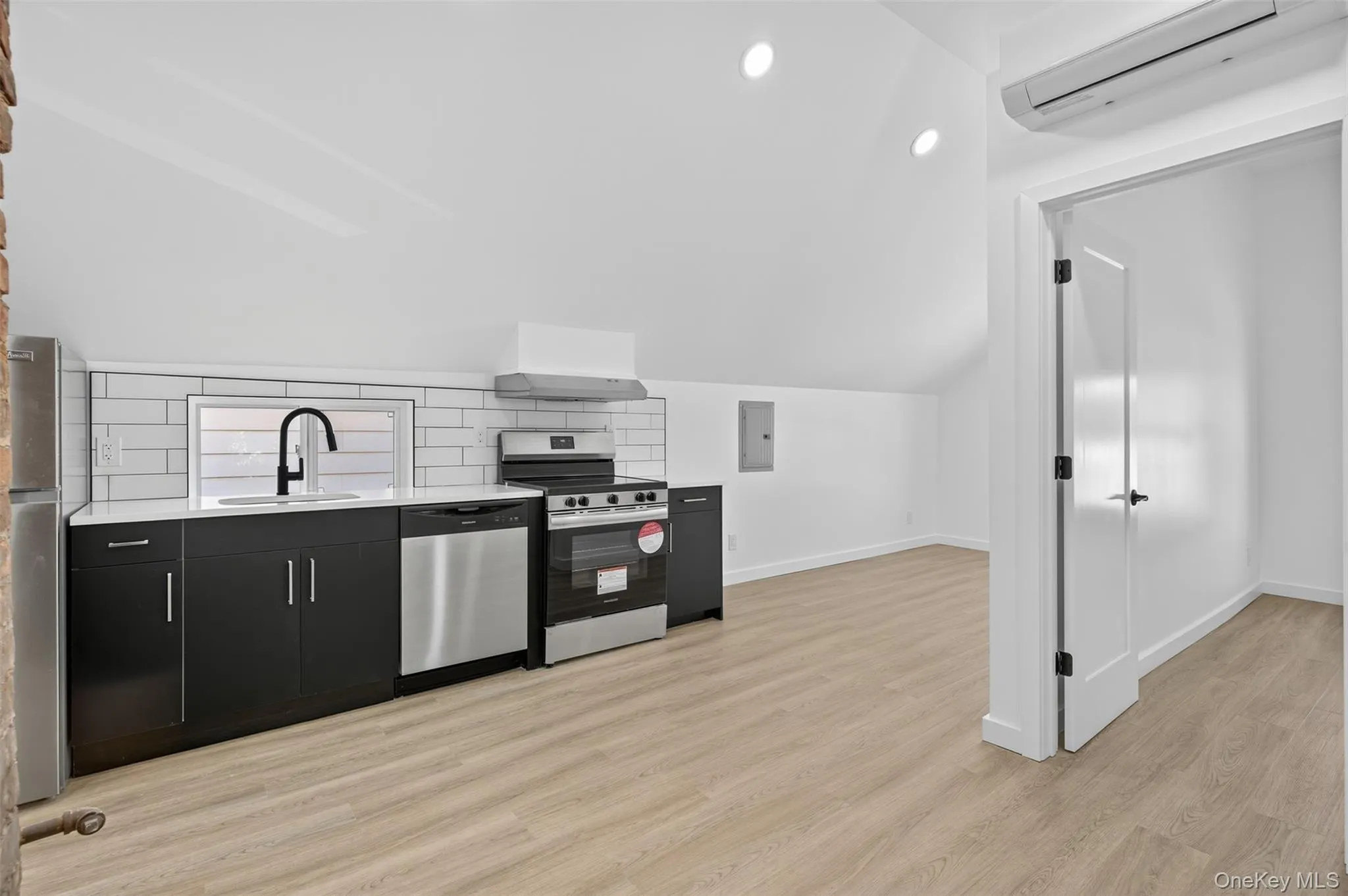 Kitchen featuring recessed lighting, light wood-type flooring, stainless steel appliances, an AC wall unit, and dark cabinets Kitchen featuring recessed lighting, light wood-type flooring, stainless steel appliances, an AC wall unit, and dark cabinets