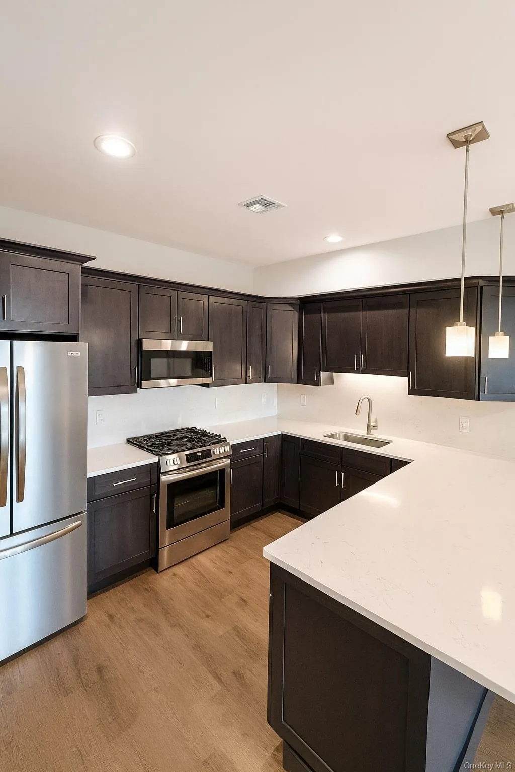 Kitchen with stainless steel appliances, decorative light fixtures, dark brown cabinetry, light wood finished floors, and light stone counters Kitchen with stainless steel appliances, decorative light fixtures, dark brown cabinetry, light wood finished floors, and light stone counters