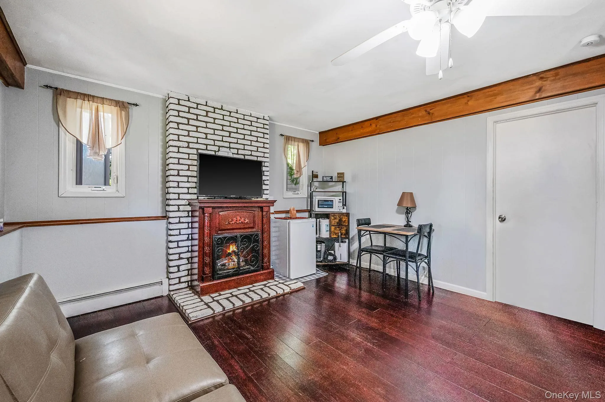 Living room featuring wood-type flooring, a baseboard radiator, beam ceiling, a fireplace, and a ceiling fan Living room featuring wood-type flooring, a baseboard radiator, beam ceiling, a fireplace, and a ceiling fan