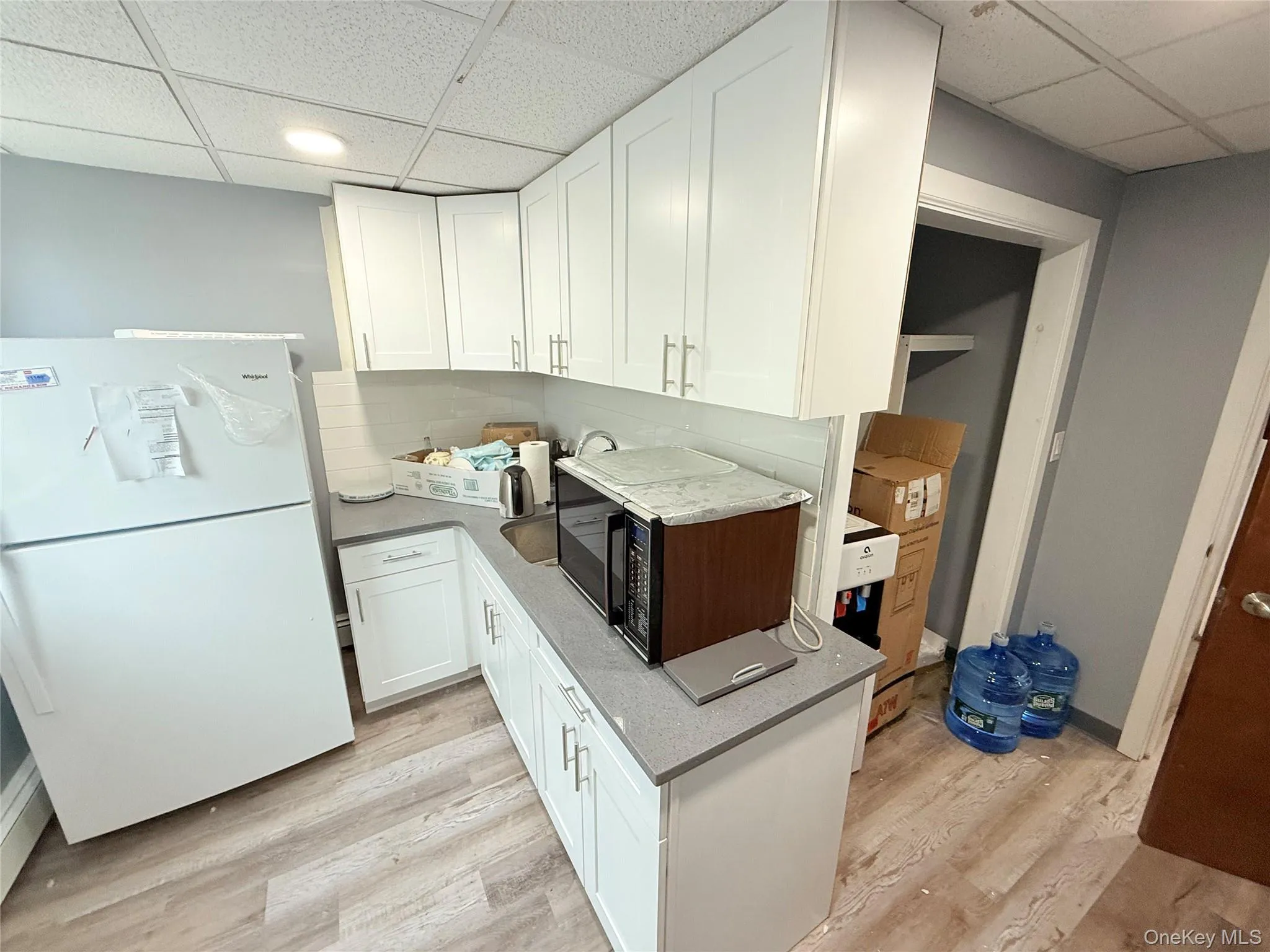 Kitchen featuring a drop ceiling, freestanding refrigerator, white cabinets, black microwave, and light wood finished floors Kitchen featuring a drop ceiling, freestanding refrigerator, white cabinets, black microwave, and light wood finished floors