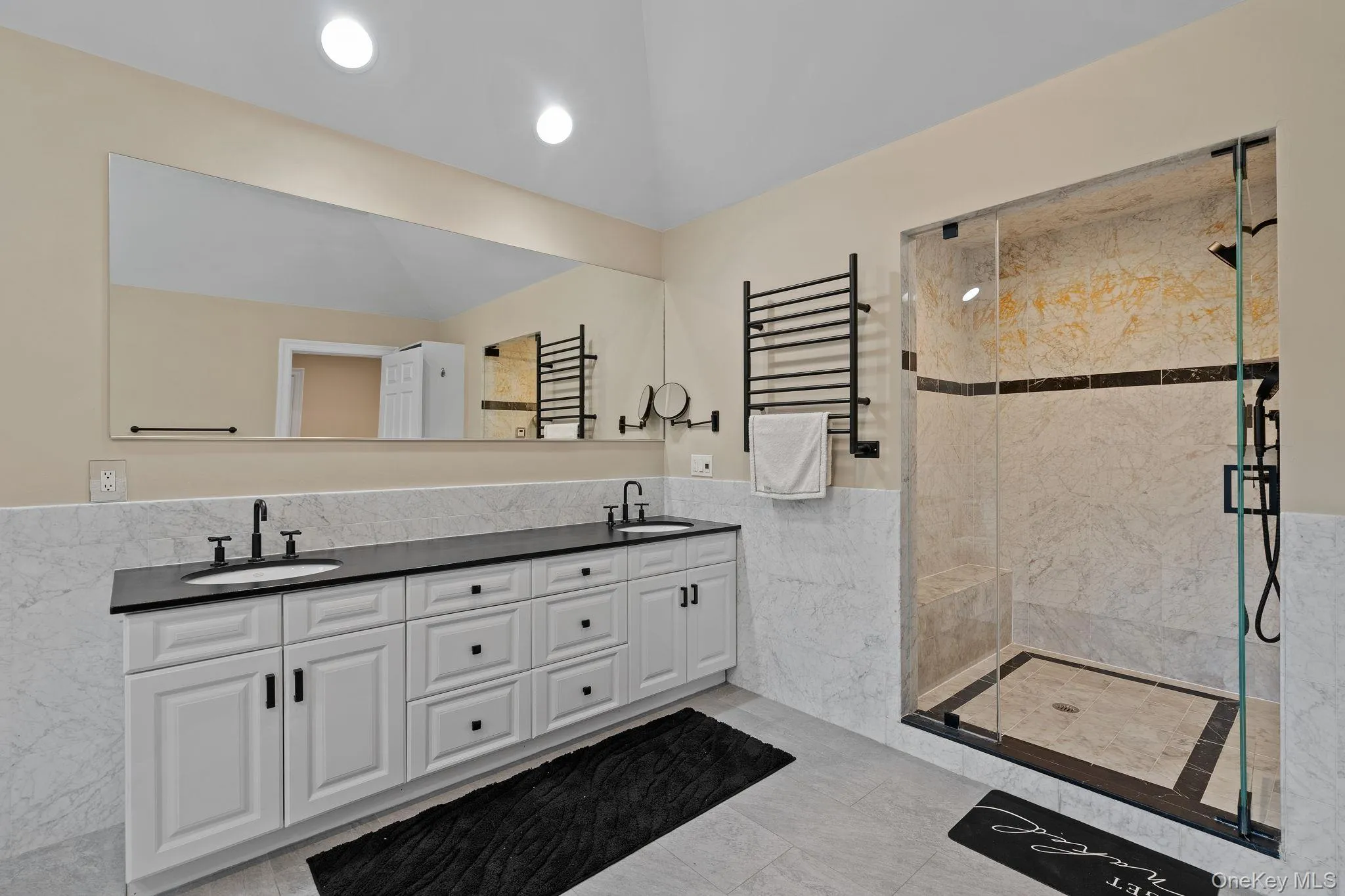 Bathroom with tile walls, a stall shower, double vanity, wainscoting, and a towel warming rack Bathroom with tile walls, a stall shower, double vanity, wainscoting, and a towel warming rack