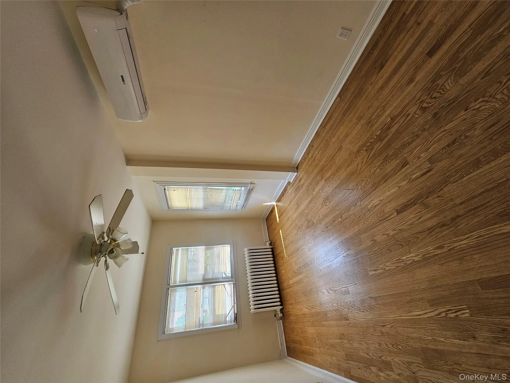 Empty room featuring radiator heating unit, a wall mounted AC, wood finished floors, and ceiling fan Empty room featuring radiator heating unit, a wall mounted AC, wood finished floors, and ceiling fan