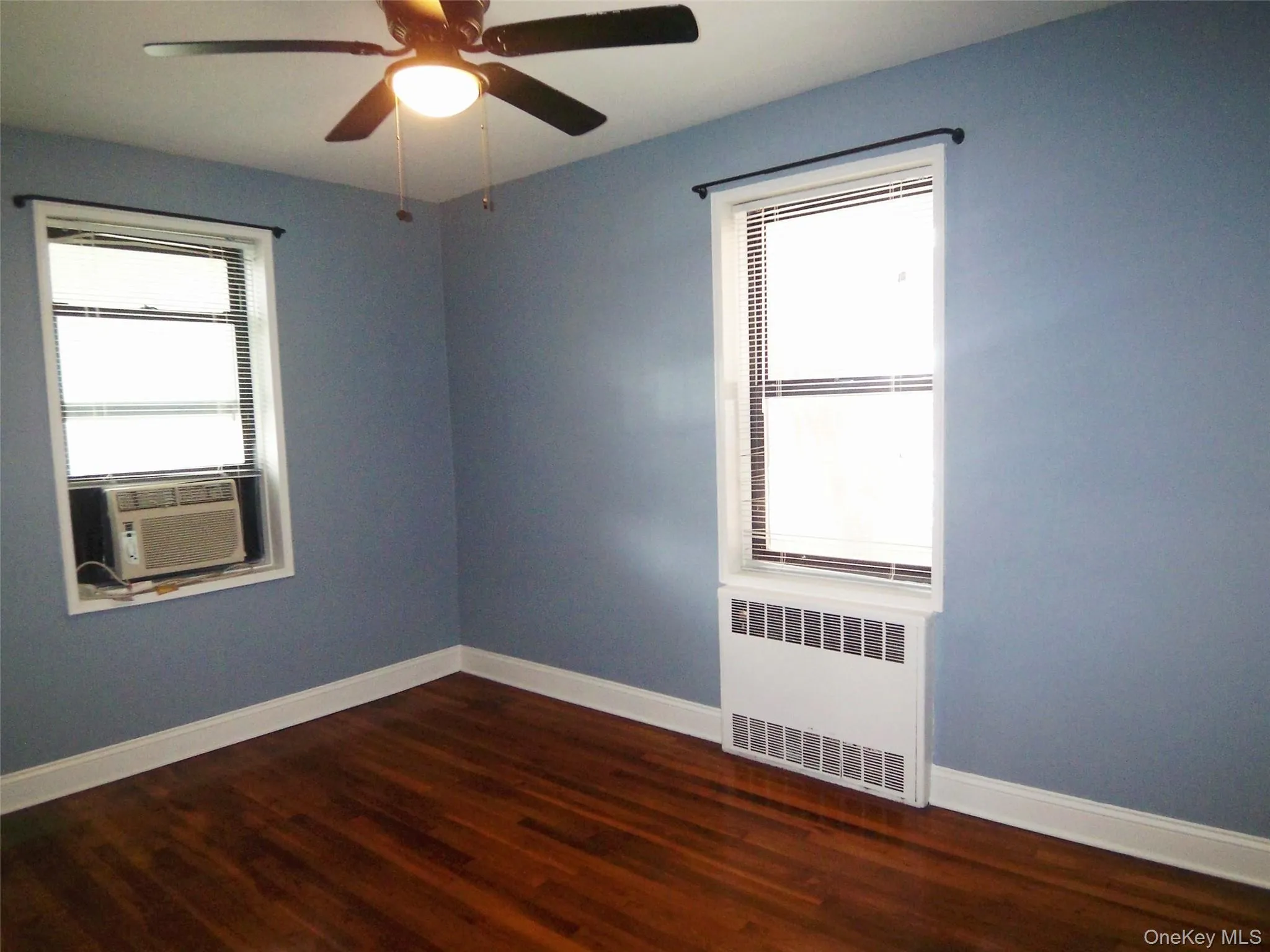 Empty room featuring dark wood-style flooring, radiator heating unit, cooling unit, and ceiling fan Empty room featuring dark wood-style flooring, radiator heating unit, cooling unit, and ceiling fan
