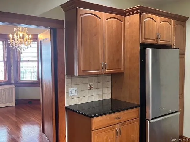 Kitchen featuring freestanding refrigerator, radiator, decorative backsplash, brown cabinets, and a chandelier Kitchen featuring freestanding refrigerator, radiator, decorative backsplash, brown cabinets, and a chandelier