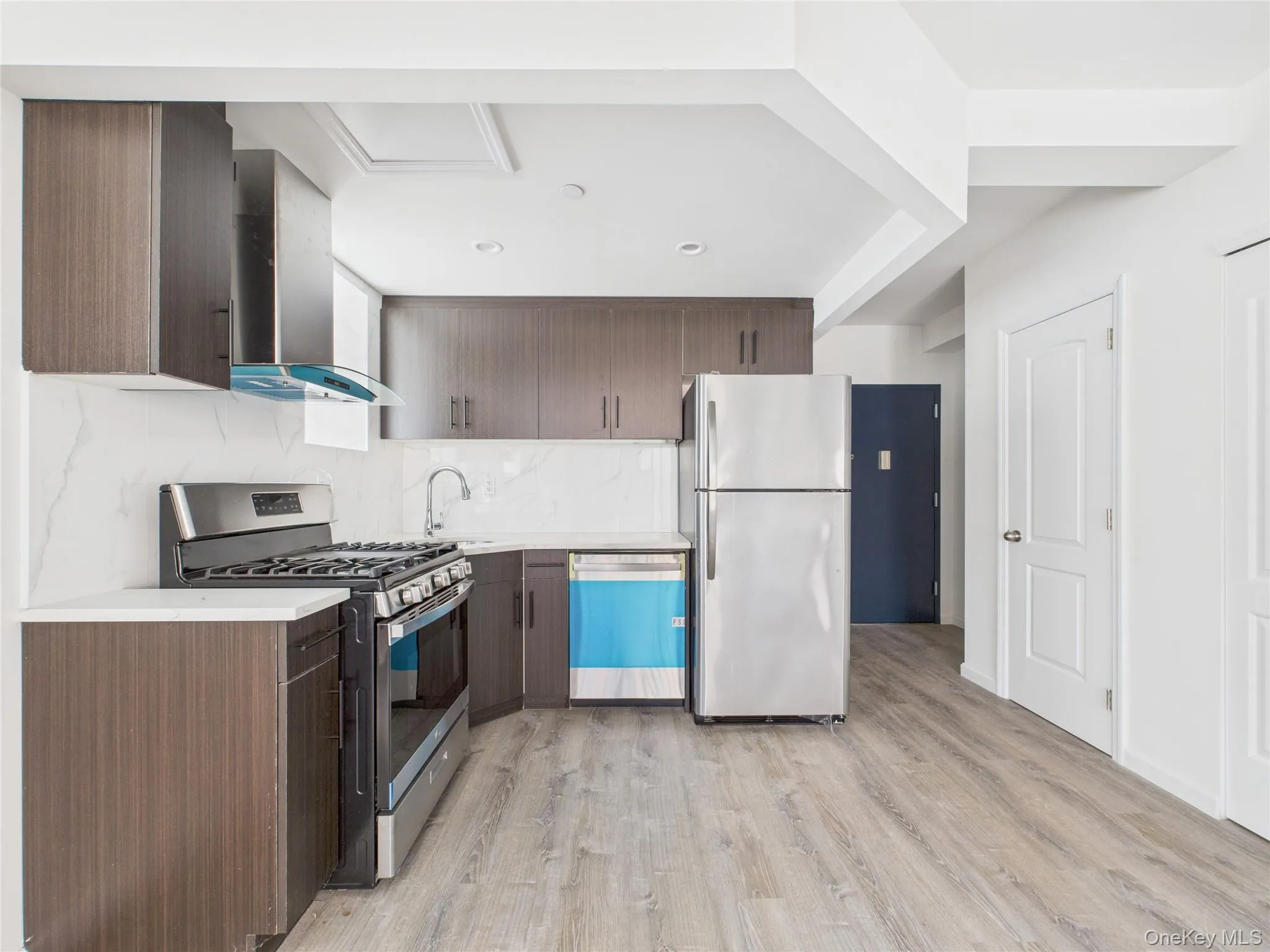 Kitchen with appliances with stainless steel finishes, tasteful backsplash, wall chimney exhaust hood, light wood-type flooring, and dark brown cabinetry Kitchen with appliances with stainless steel finishes, tasteful backsplash, wall chimney exhaust hood, light wood-type flooring, and dark brown cabinetry