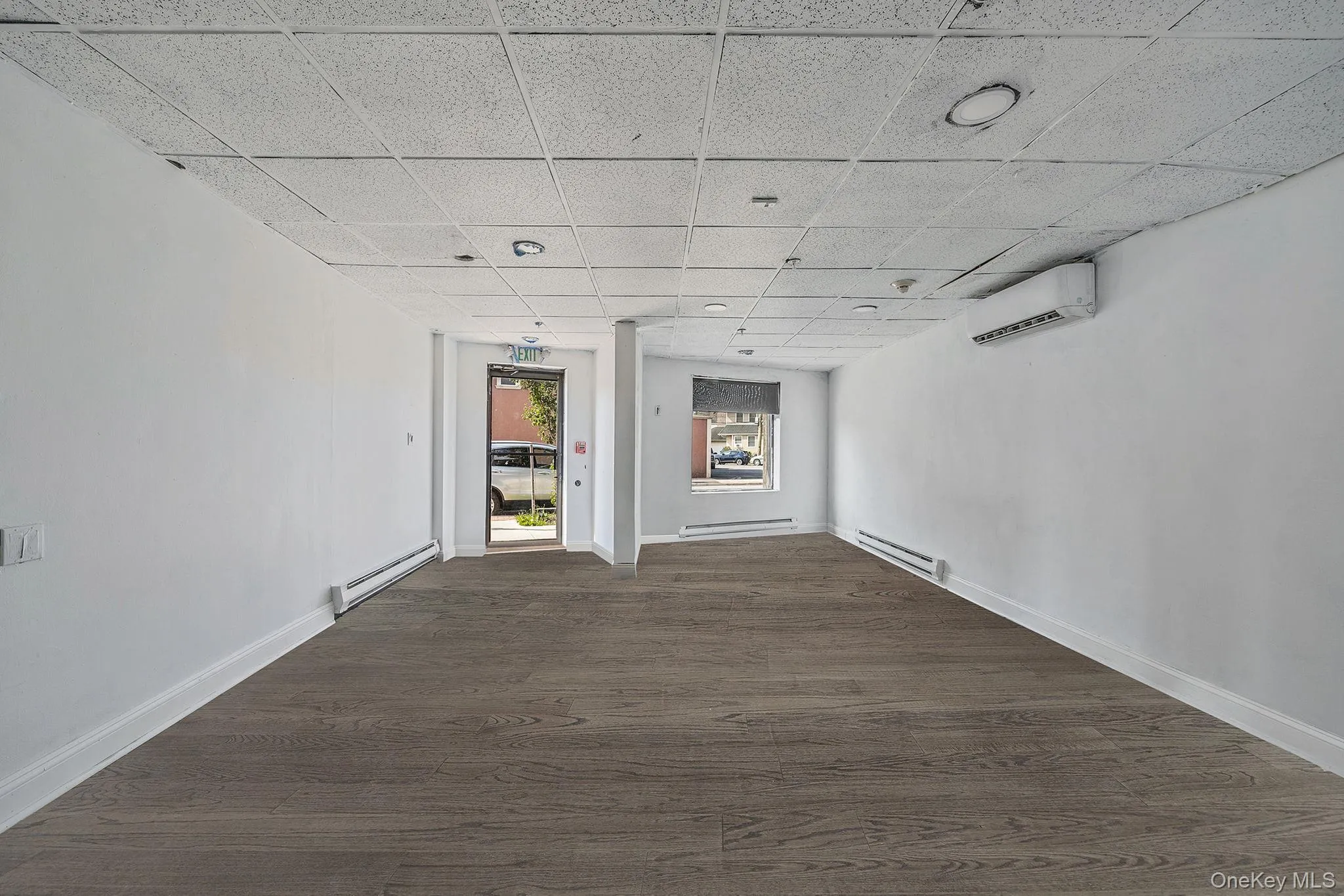 Empty room with a drop ceiling, dark wood-style flooring, baseboard heating, an AC wall unit, and a baseboard heating unit Empty room with a drop ceiling, dark wood-style flooring, baseboard heating, an AC wall unit, and a baseboard heating unit