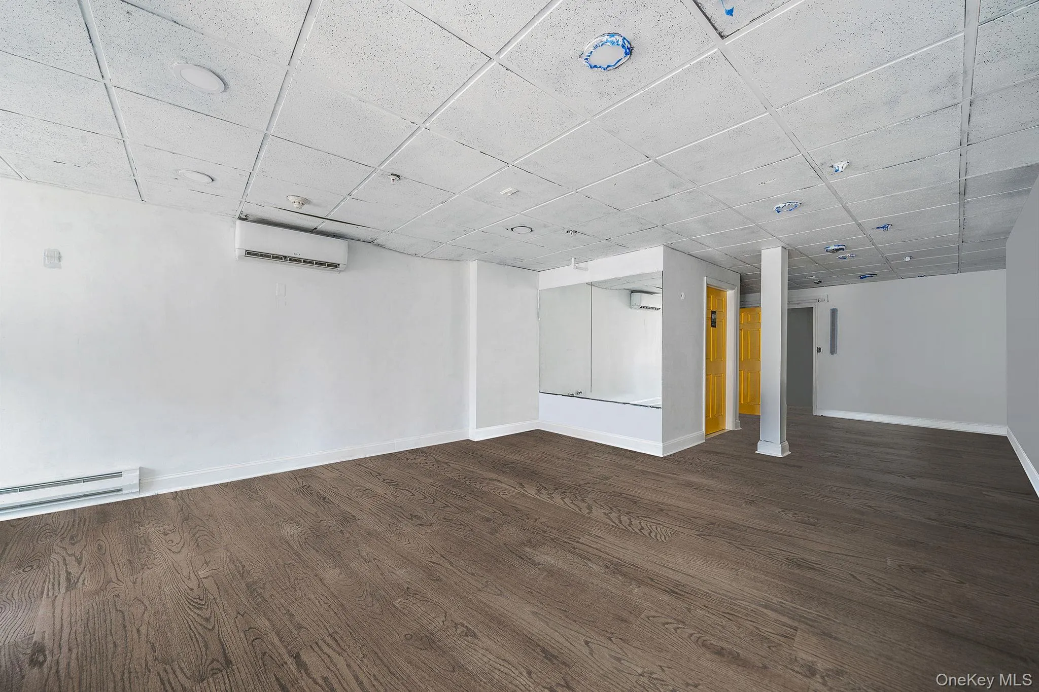 Below grade area featuring a drop ceiling, dark wood-style floors, a baseboard radiator, and a wall unit AC Below grade area featuring a drop ceiling, dark wood-style floors, a baseboard radiator, and a wall unit AC