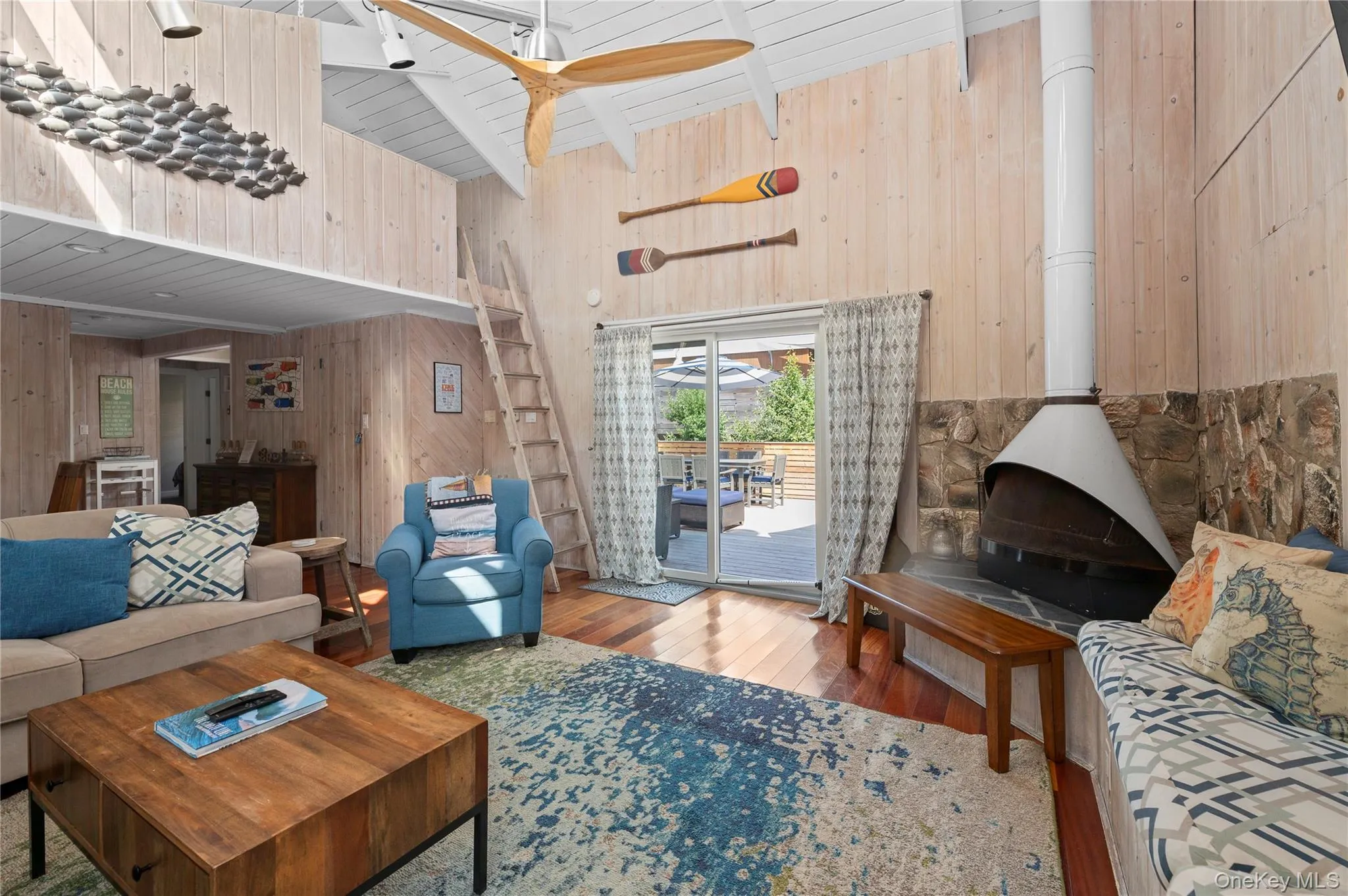 Living room featuring wood-type flooring, high vaulted ceiling, a wood stove, wood walls, and a ceiling fan Living room featuring wood-type flooring, high vaulted ceiling, a wood stove, wood walls, and a ceiling fan