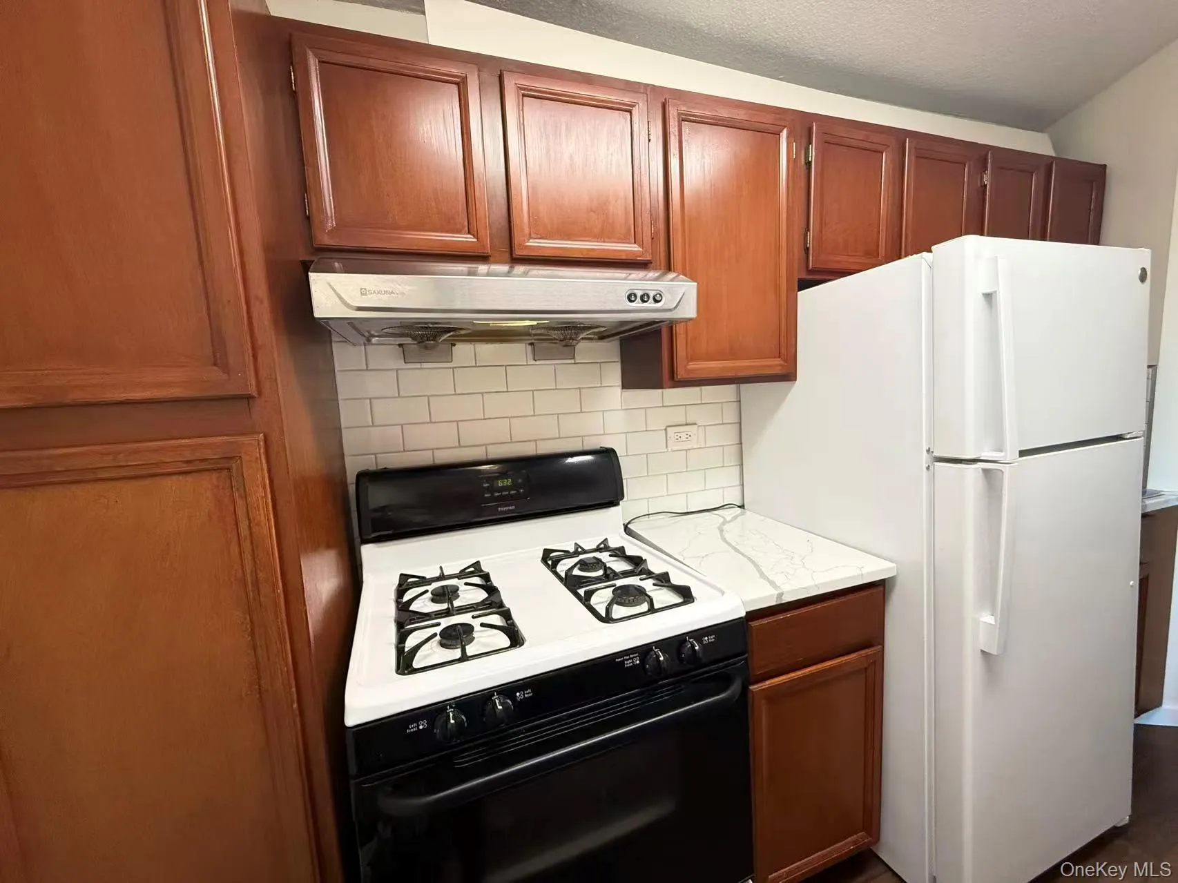 Kitchen featuring gas range, decorative backsplash, freestanding refrigerator, under cabinet range hood, and brown cabinetry Kitchen featuring gas range, decorative backsplash, freestanding refrigerator, under cabinet range hood, and brown cabinetry