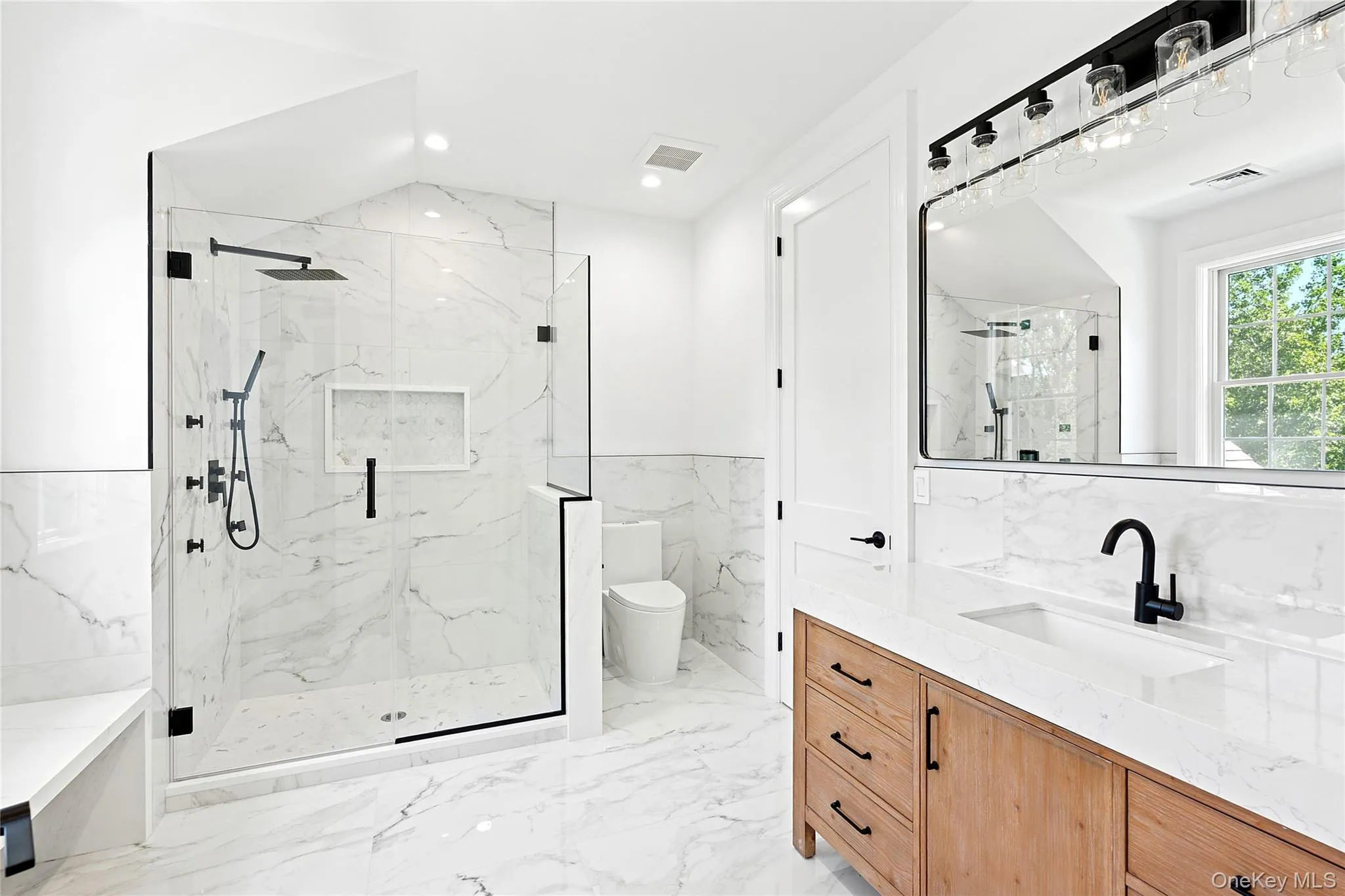 Full bathroom featuring vanity, a marble finish shower, tile walls, light marble finish flooring, and wainscoting Full bathroom featuring vanity, a marble finish shower, tile walls, light marble finish flooring, and wainscoting