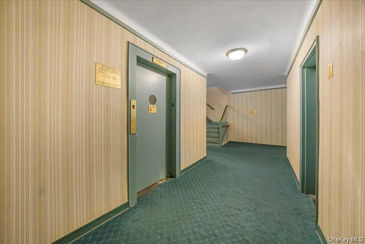 Corridor featuring stairway, elevator, wallpapered walls, and dark carpet Corridor featuring stairway, elevator, wallpapered walls, and dark carpet