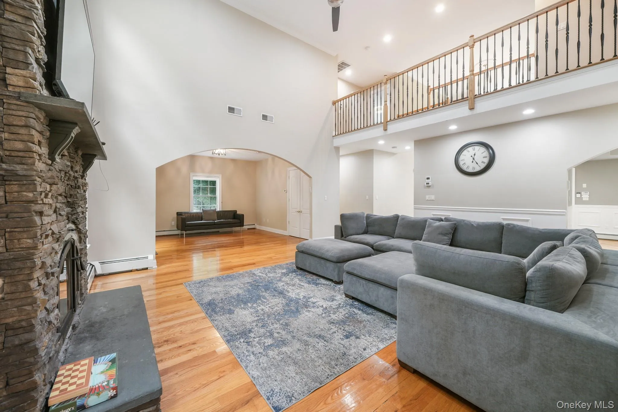 Living area featuring arched walkways, light wood finished floors, a stone fireplace, a towering ceiling, and a baseboard radiator Living area featuring arched walkways, light wood finished floors, a stone fireplace, a towering ceiling, and a baseboard radiator