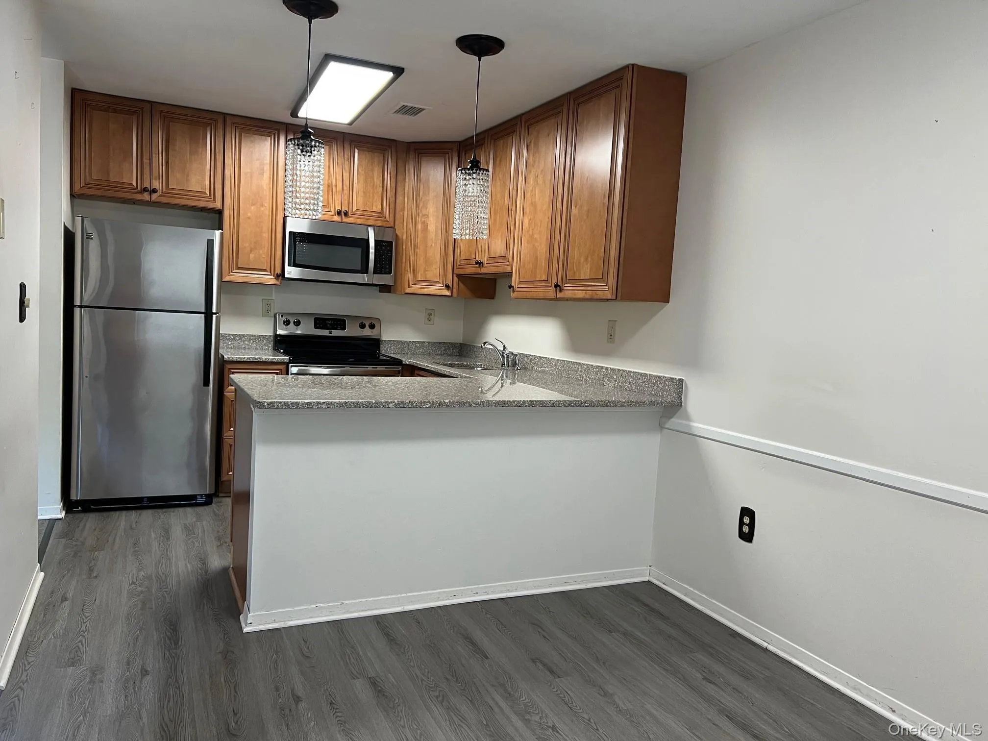 Kitchen featuring appliances with stainless steel finishes, dark wood finished floors, hanging light fixtures, light stone countertops, and brown cabinets Kitchen featuring appliances with stainless steel finishes, dark wood finished floors, hanging light fixtures, light stone countertops, and brown cabinets