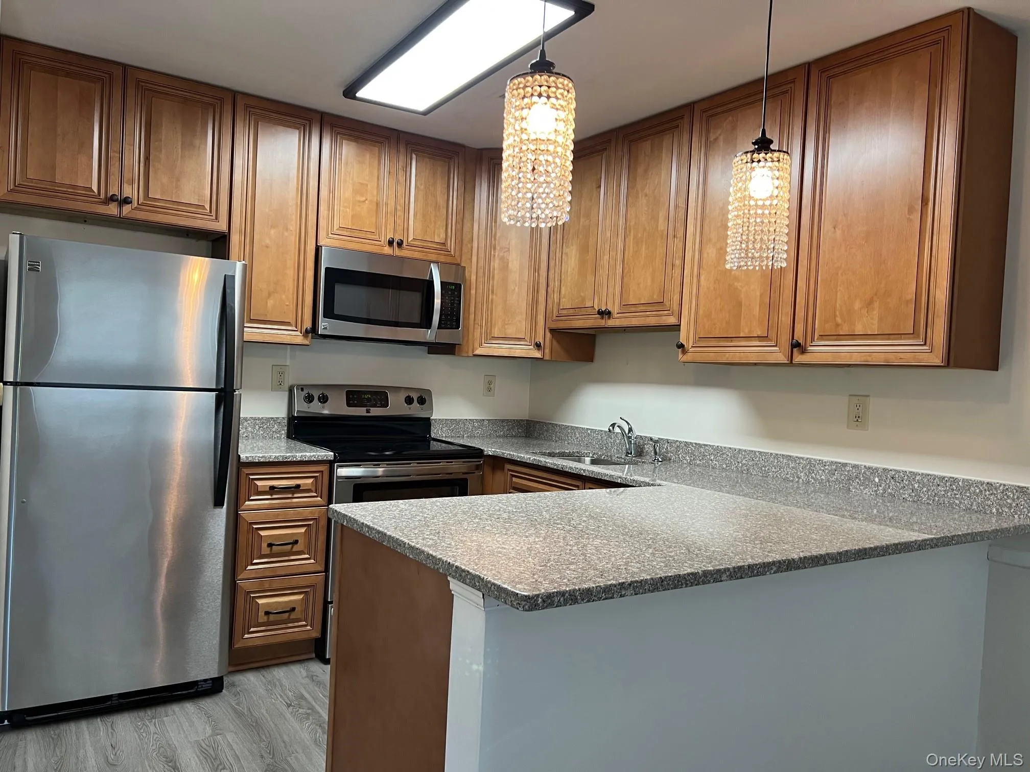Kitchen with stainless steel appliances, brown cabinets, decorative light fixtures, light wood-type flooring, and dark stone countertops Kitchen with stainless steel appliances, brown cabinets, decorative light fixtures, light wood-type flooring, and dark stone countertops