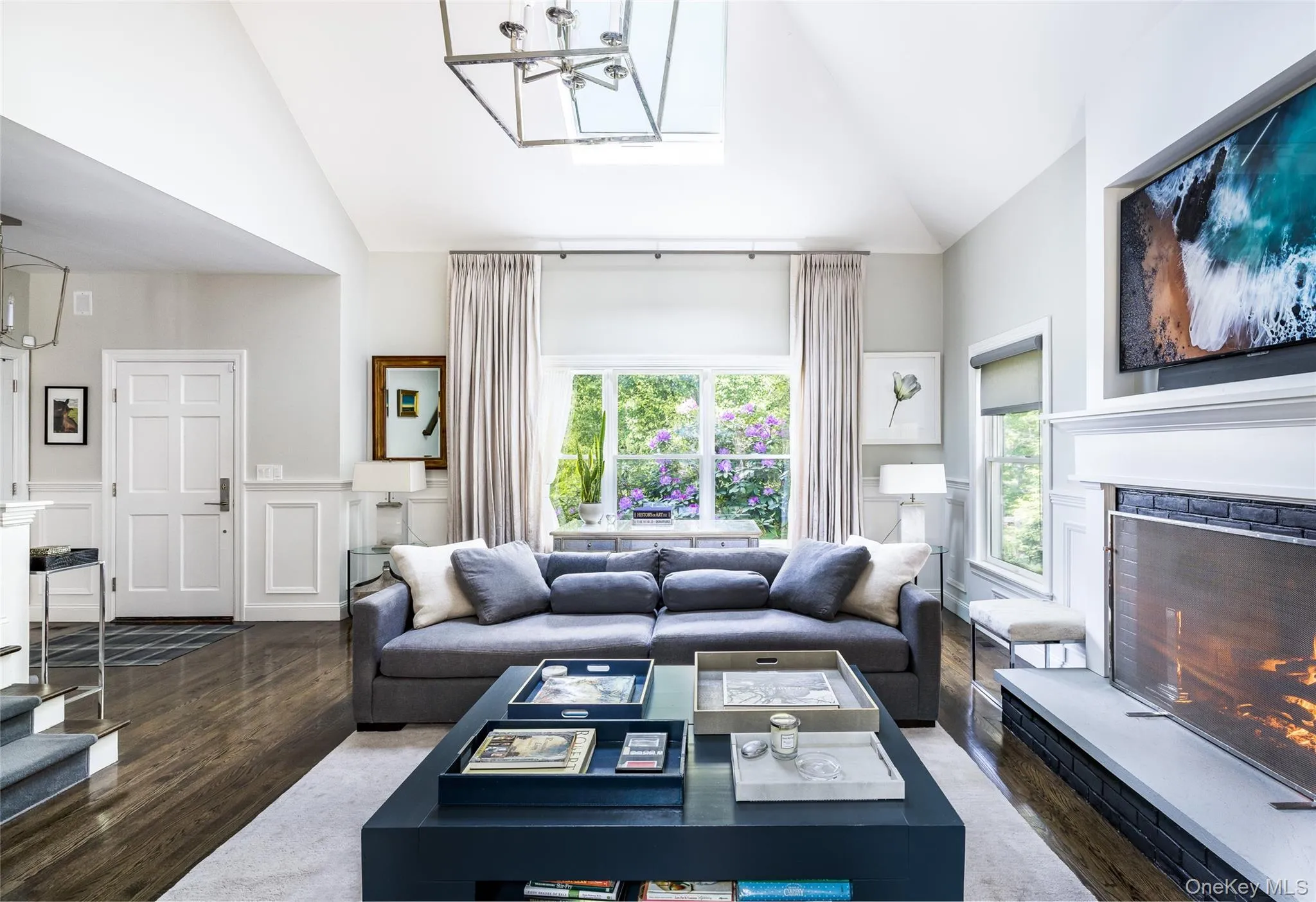 Living room with a lit fireplace, a wainscoted wall, a decorative wall, wood finished floors, and high vaulted ceiling Living room with a lit fireplace, a wainscoted wall, a decorative wall, wood finished floors, and high vaulted ceiling