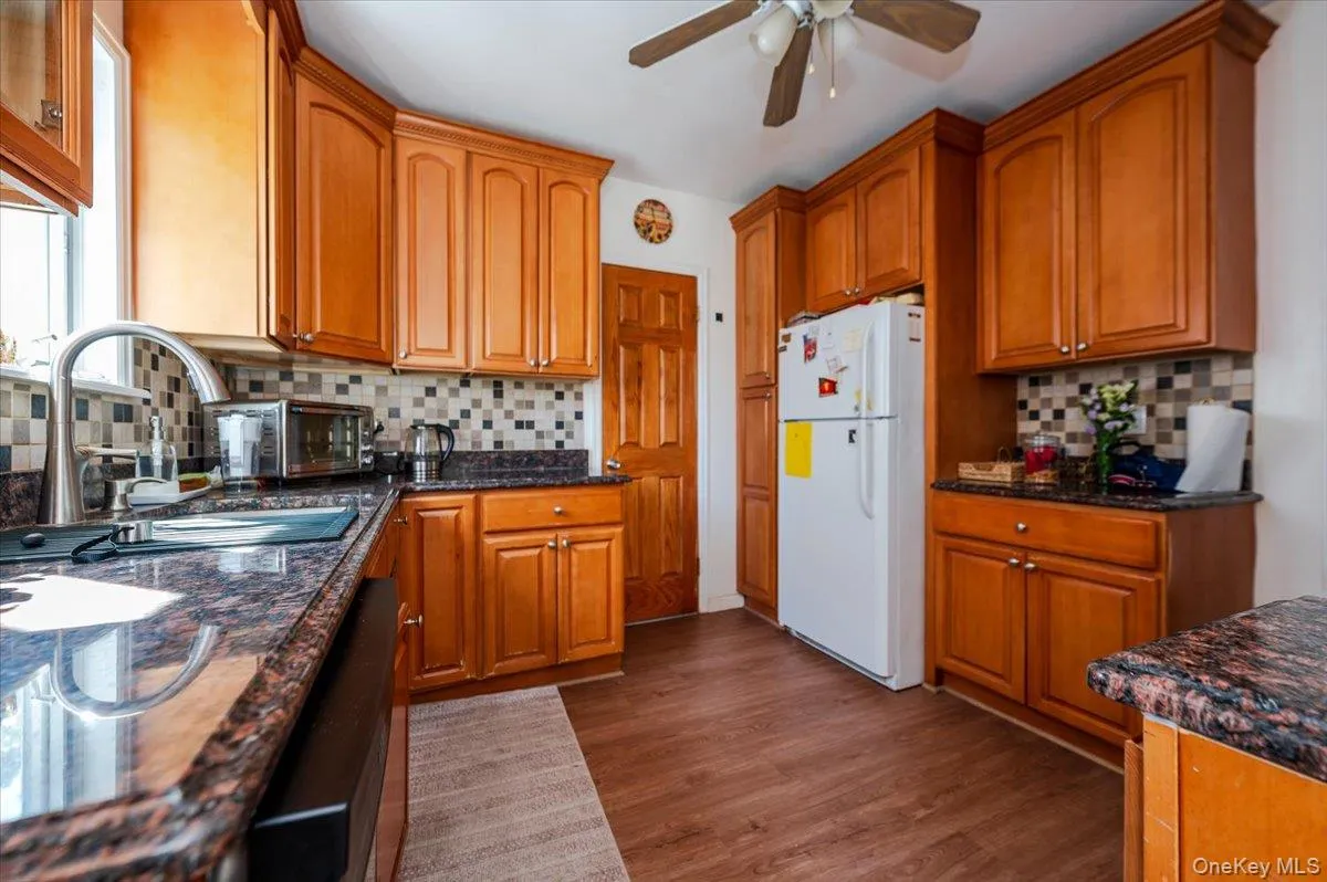 Kitchen featuring decorative backsplash, freestanding refrigerator, dark stone counters, and brown cabinets Kitchen featuring decorative backsplash, freestanding refrigerator, dark stone counters, and brown cabinets