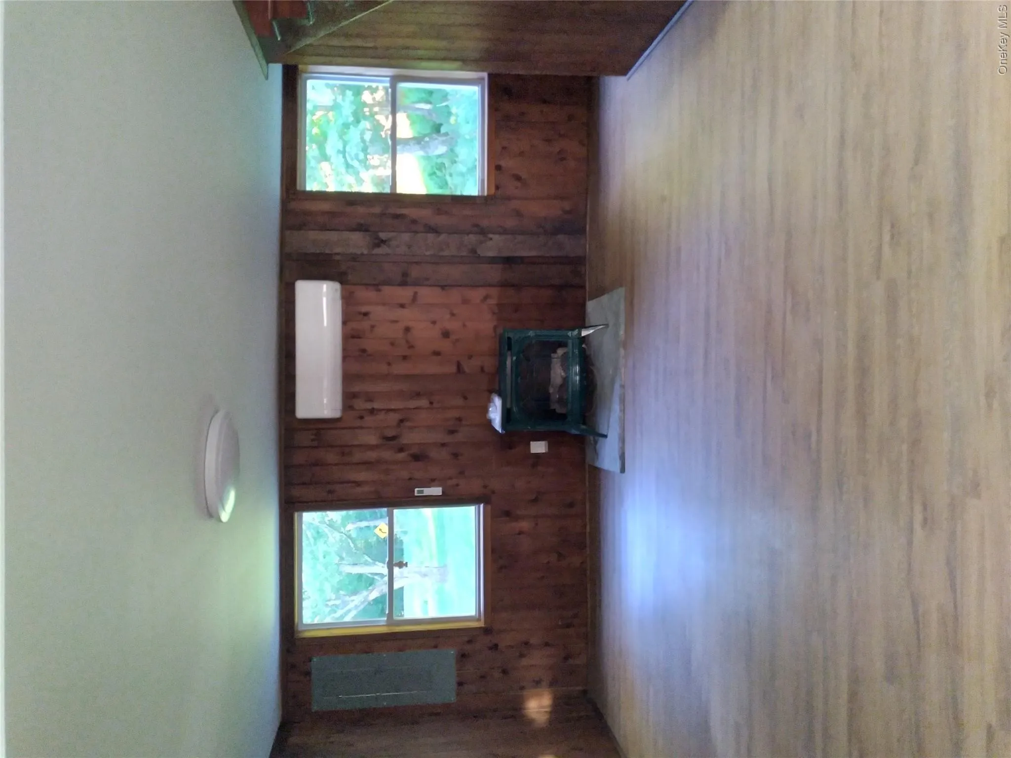 Unfurnished living room featuring a wood stove, wood walls, light wood finished floors, a wall mounted AC, and electric panel Unfurnished living room featuring a wood stove, wood walls, light wood finished floors, a wall mounted AC, and electric panel