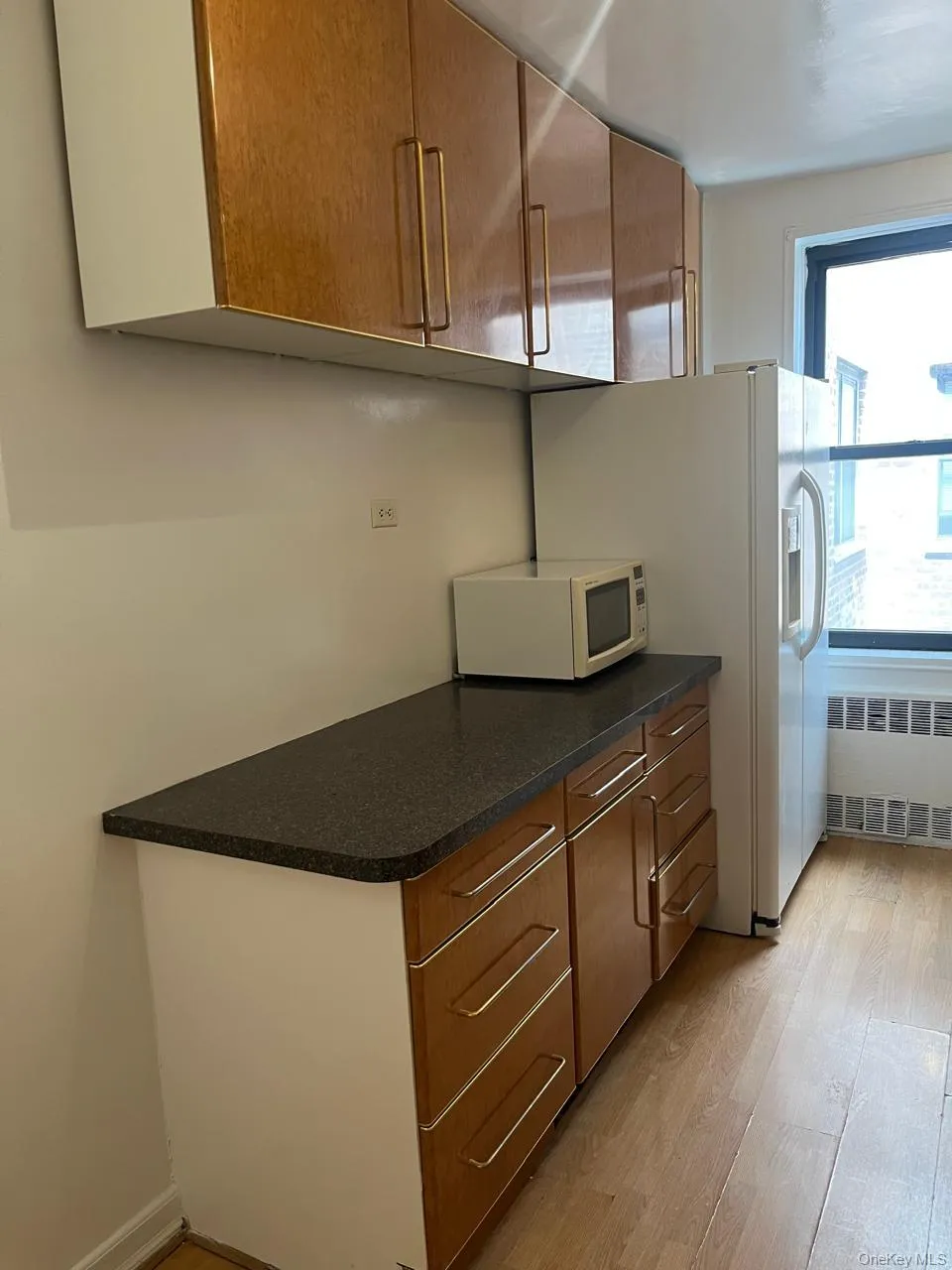 Kitchen with brown cabinets, dark countertops, light wood finished floors, radiator heating unit, and white microwave Kitchen with brown cabinets, dark countertops, light wood finished floors, radiator heating unit, and white microwave
