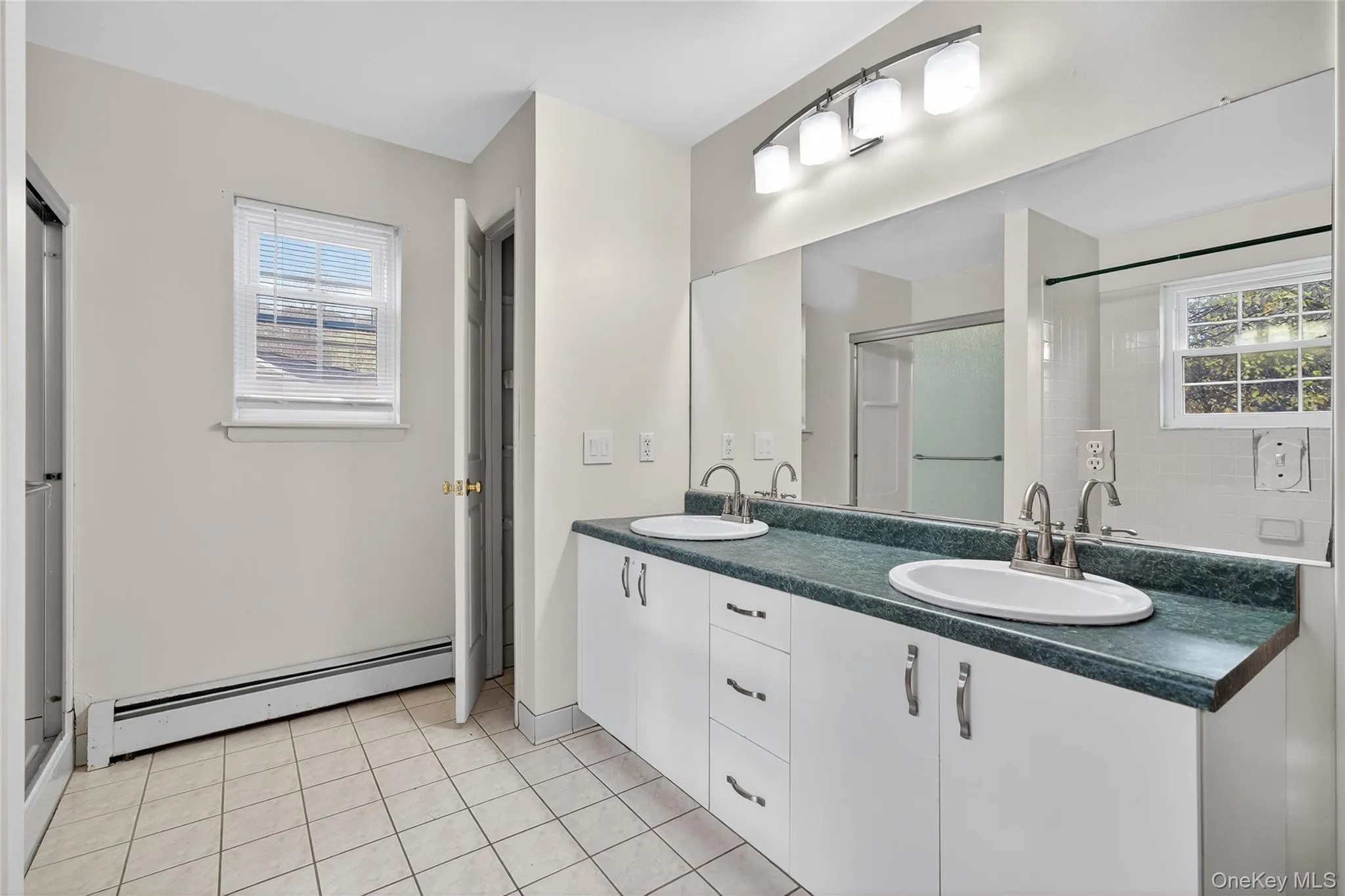 Bathroom featuring baseboard heating, double vanity, light tile patterned floors, and a shower stall Bathroom featuring baseboard heating, double vanity, light tile patterned floors, and a shower stall