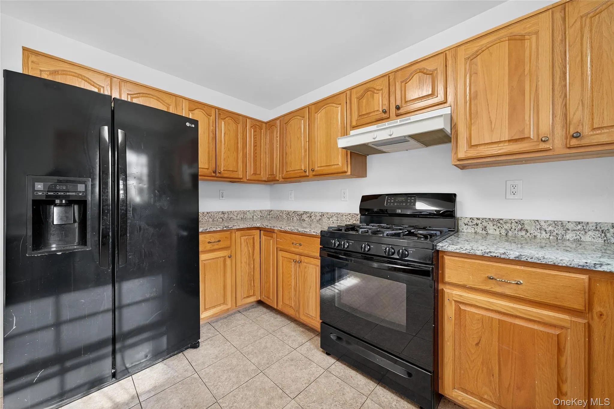Kitchen featuring black appliances, light stone counters, under cabinet range hood, light tile patterned flooring, and brown cabinetry Kitchen featuring black appliances, light stone counters, under cabinet range hood, light tile patterned flooring, and brown cabinetry