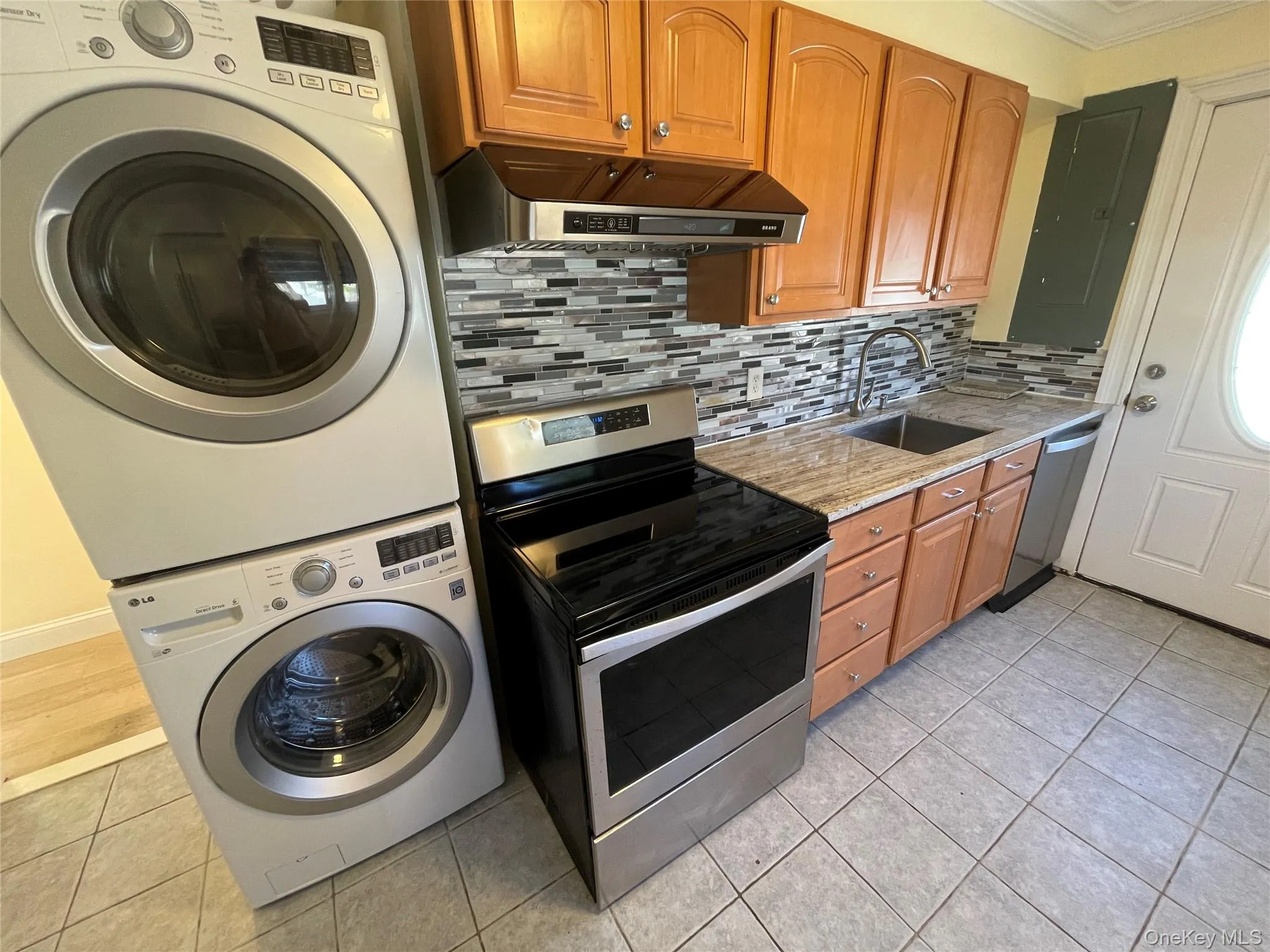 Kitchen featuring appliances with stainless steel finishes, light tile patterned floors, exhaust hood, backsplash, and brown cabinetry Kitchen featuring appliances with stainless steel finishes, light tile patterned floors, exhaust hood, backsplash, and brown cabinetry