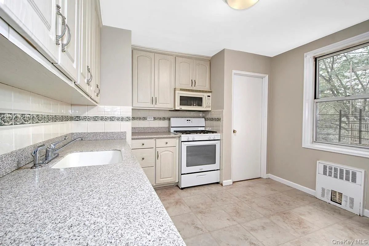 Kitchen featuring white appliances, decorative backsplash, radiator, light stone counters, and light tile patterned floors Kitchen featuring white appliances, decorative backsplash, radiator, light stone counters, and light tile patterned floors