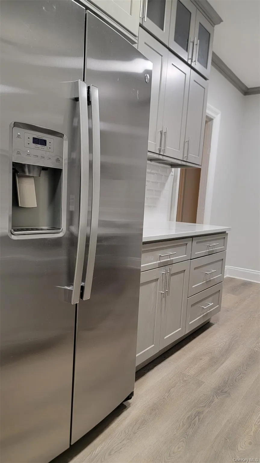 Kitchen with stainless steel refrigerator with ice dispenser, light wood-style floors, ornamental molding, gray cabinetry, and glass insert cabinets Kitchen with stainless steel refrigerator with ice dispenser, light wood-style floors, ornamental molding, gray cabinetry, and glass insert cabinets