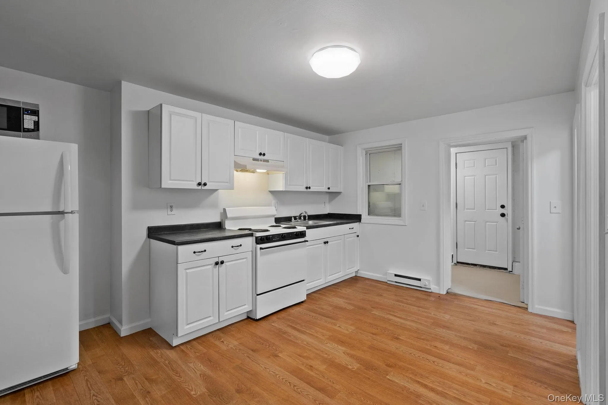 Kitchen featuring white appliances, white cabinetry, dark countertops, light wood-style flooring, and under cabinet range hood Kitchen featuring white appliances, white cabinetry, dark countertops, light wood-style flooring, and under cabinet range hood