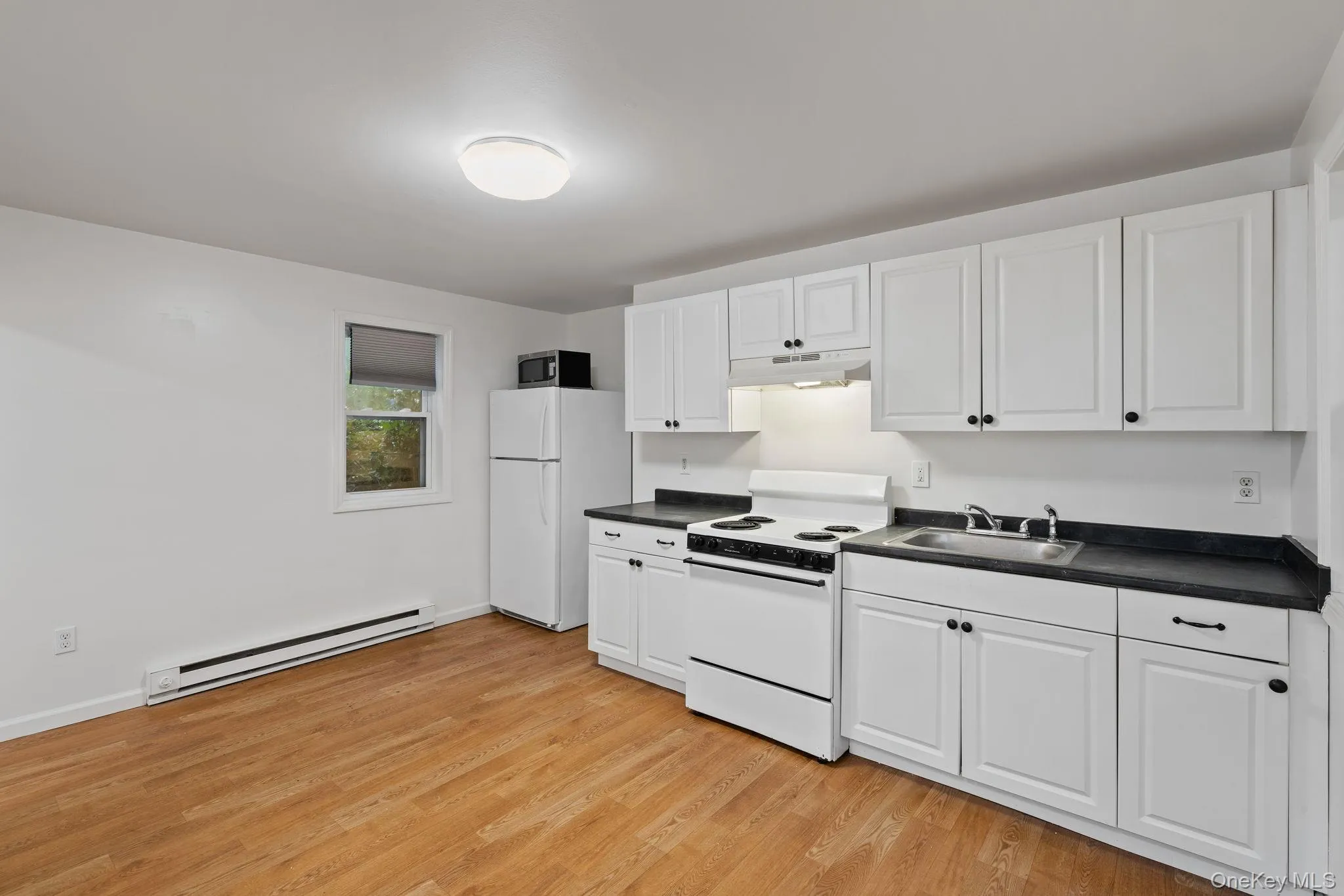 Kitchen featuring white appliances, dark countertops, white cabinets, and a baseboard heating unit Kitchen featuring white appliances, dark countertops, white cabinets, and a baseboard heating unit