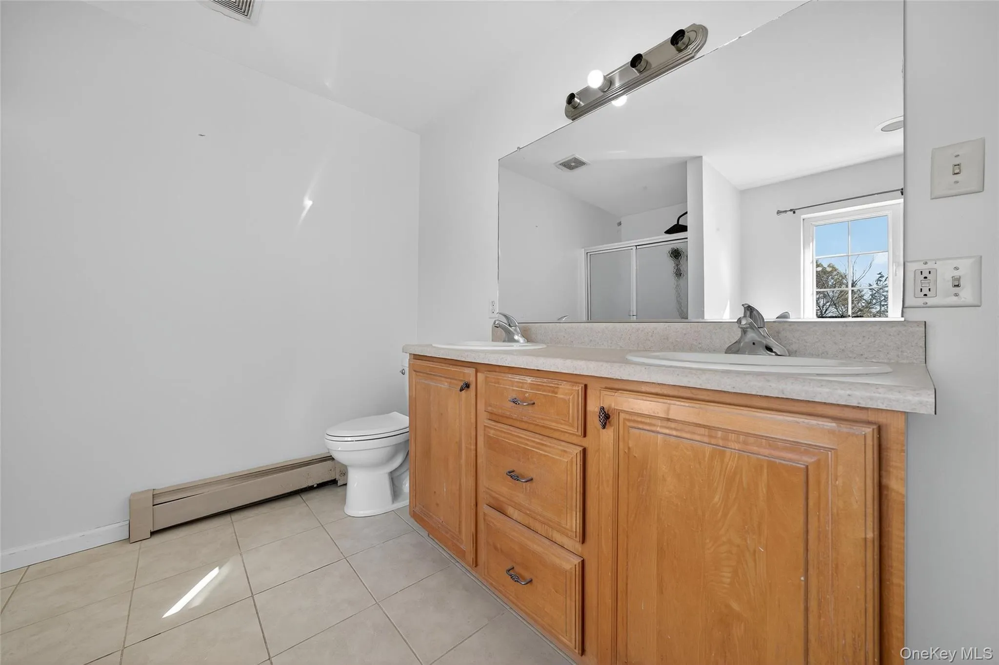 Bathroom featuring a baseboard radiator, light tile patterned flooring, double vanity, and a stall shower Bathroom featuring a baseboard radiator, light tile patterned flooring, double vanity, and a stall shower