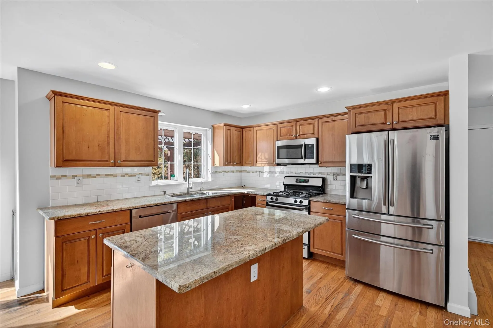 Kitchen with appliances with stainless steel finishes, light stone countertops, light wood-style flooring, brown cabinetry, and recessed lighting Kitchen with appliances with stainless steel finishes, light stone countertops, light wood-style flooring, brown cabinetry, and recessed lighting