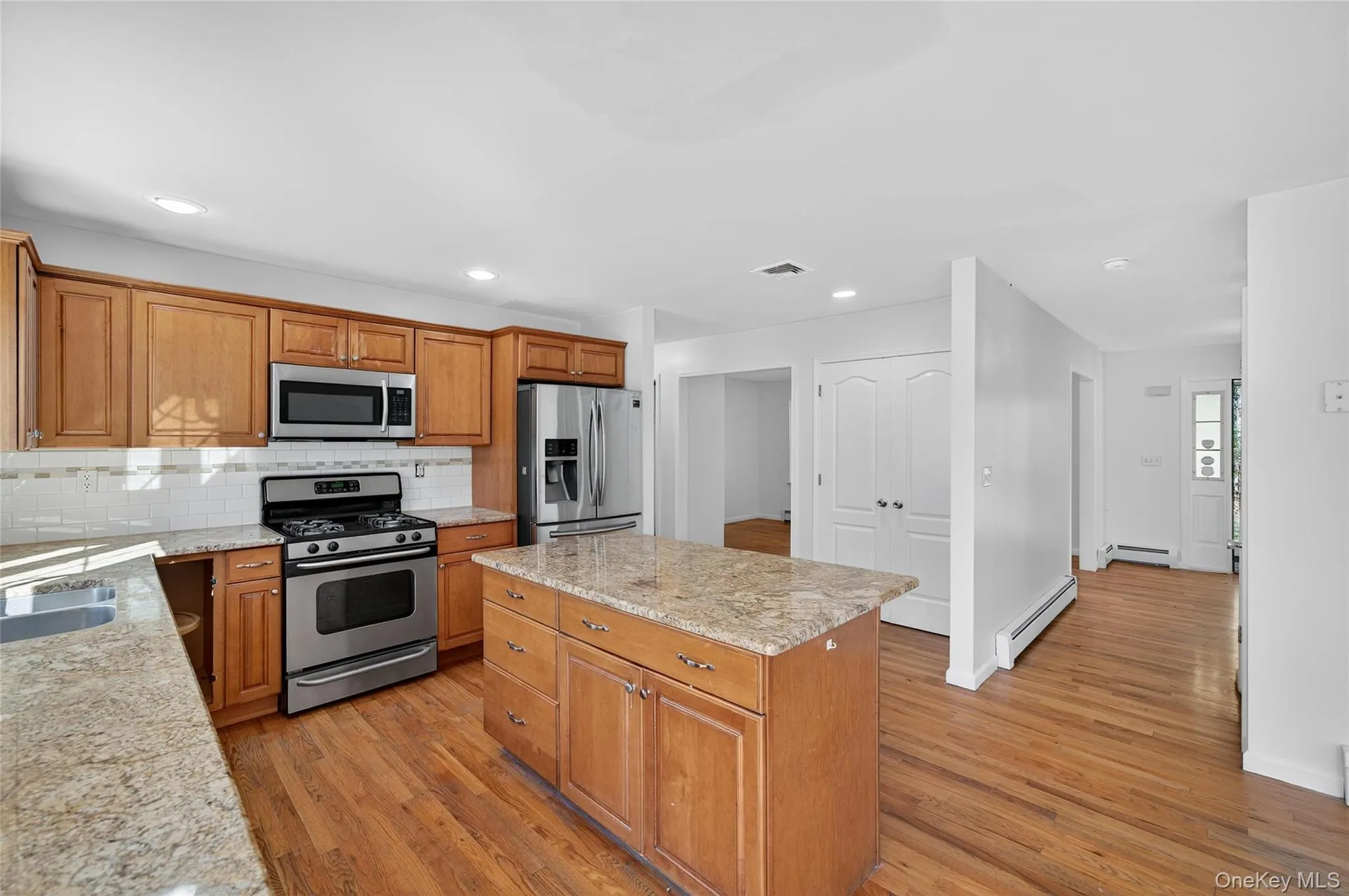 Kitchen featuring stainless steel appliances, tasteful backsplash, brown cabinetry, light stone countertops, and light wood finished floors Kitchen featuring stainless steel appliances, tasteful backsplash, brown cabinetry, light stone countertops, and light wood finished floors