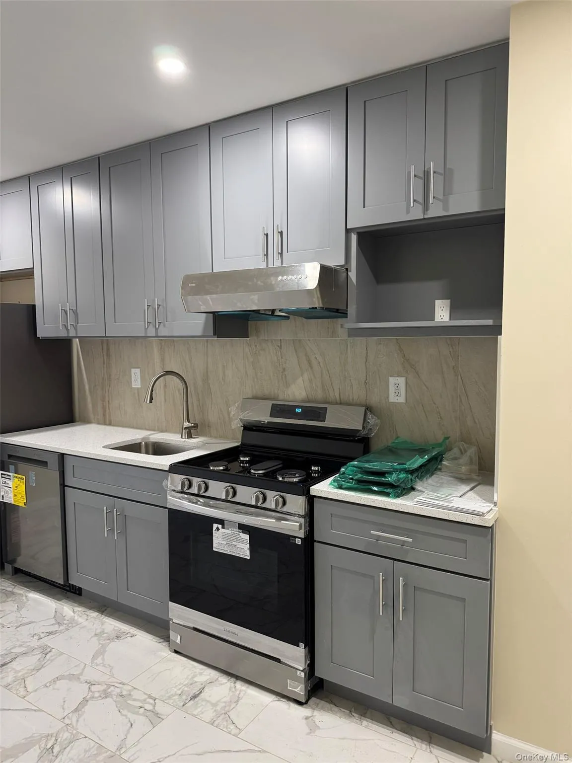 Kitchen featuring gray cabinetry, appliances with stainless steel finishes, light marble finish floors, under cabinet range hood, and decorative backsplash Kitchen featuring gray cabinetry, appliances with stainless steel finishes, light marble finish floors, under cabinet range hood, and decorative backsplash