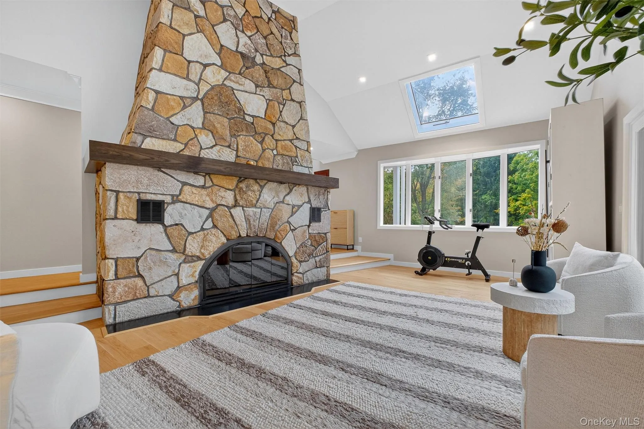 Living room with a stone wood burning fireplace, light wood floors, a skylight, high vaulted ceiling, and recessed lighting Living room with a stone wood burning fireplace, light wood floors, a skylight, high vaulted ceiling, and recessed lighting