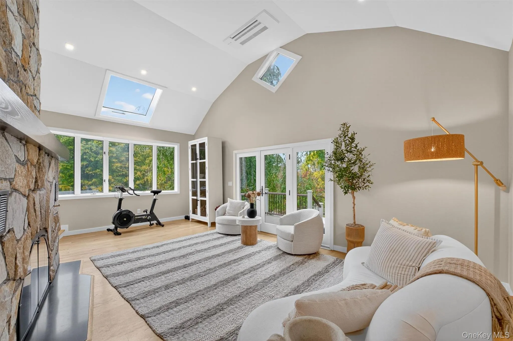 Living area featuring french doors to a deck, a fireplace, high vaulted ceiling, a skylight, and light wood flooring Living area featuring french doors to a deck, a fireplace, high vaulted ceiling, a skylight, and light wood flooring