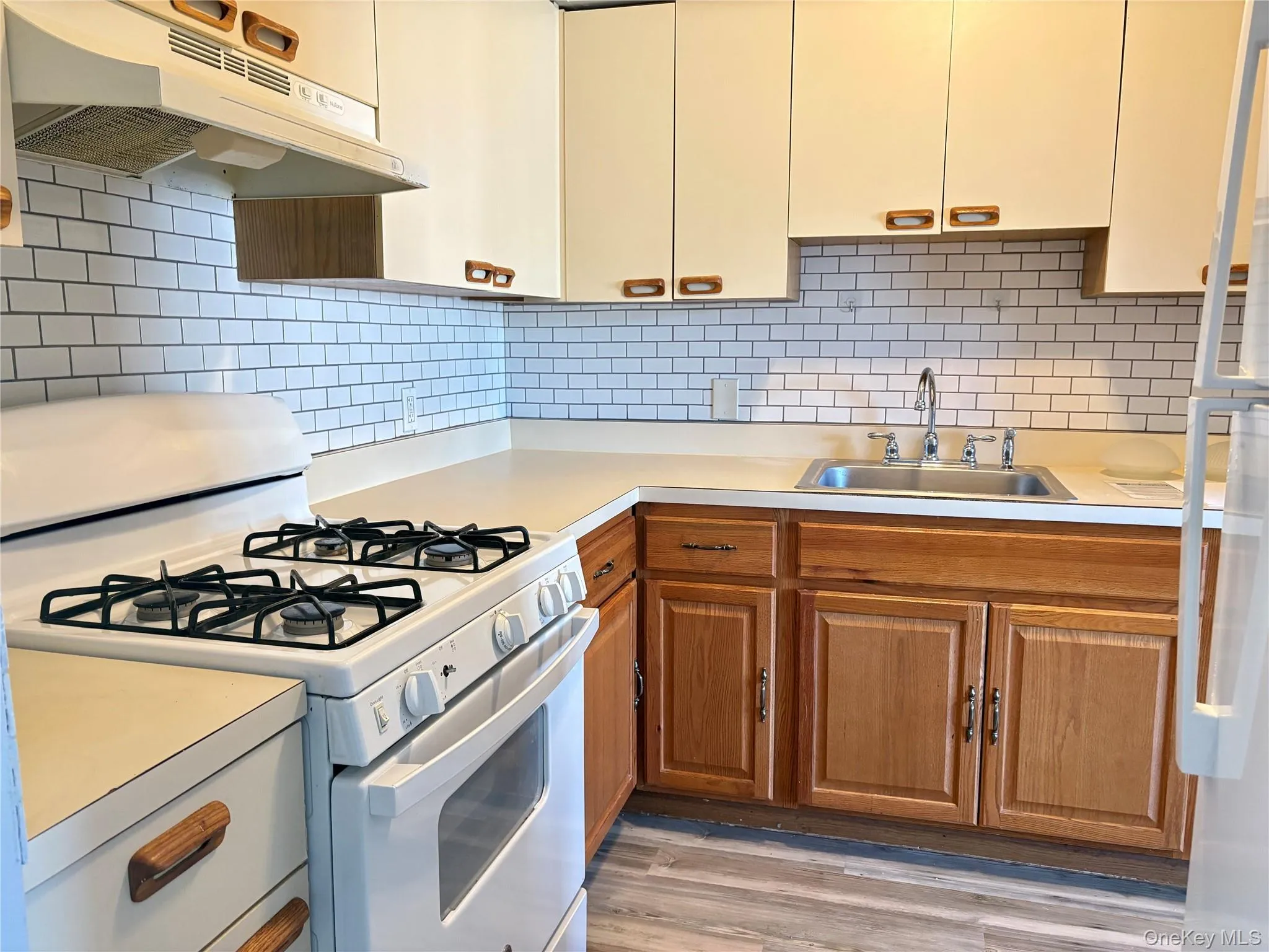 Kitchen featuring white appliances, under cabinet range hood, light countertops, backsplash, and light wood-style flooring Kitchen featuring white appliances, under cabinet range hood, light countertops, backsplash, and light wood-style flooring