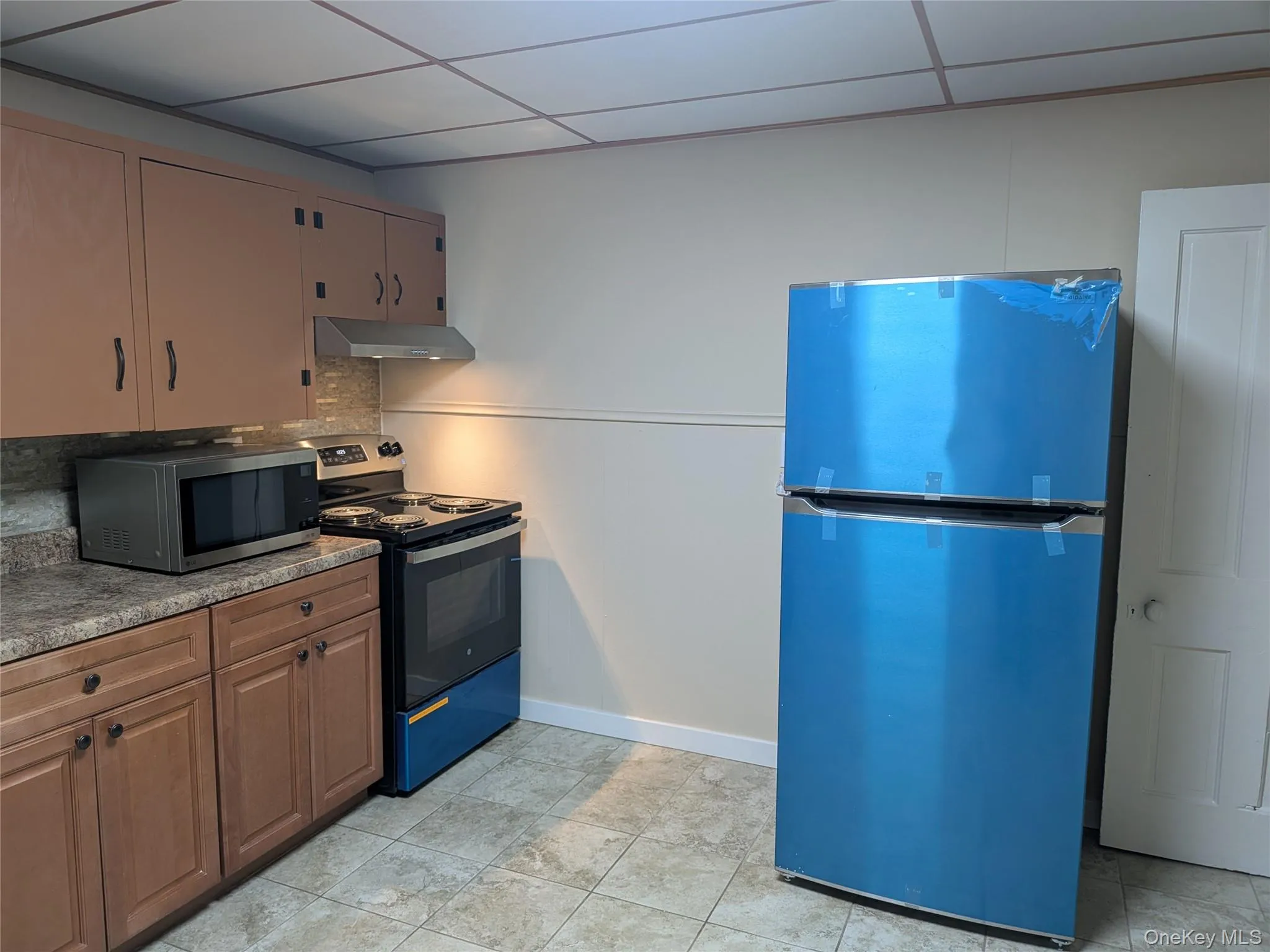 Kitchen featuring freestanding refrigerator, range with electric stovetop, stainless steel microwave, a drop ceiling, and under cabinet range hood Kitchen featuring freestanding refrigerator, range with electric stovetop, stainless steel microwave, a drop ceiling, and under cabinet range hood