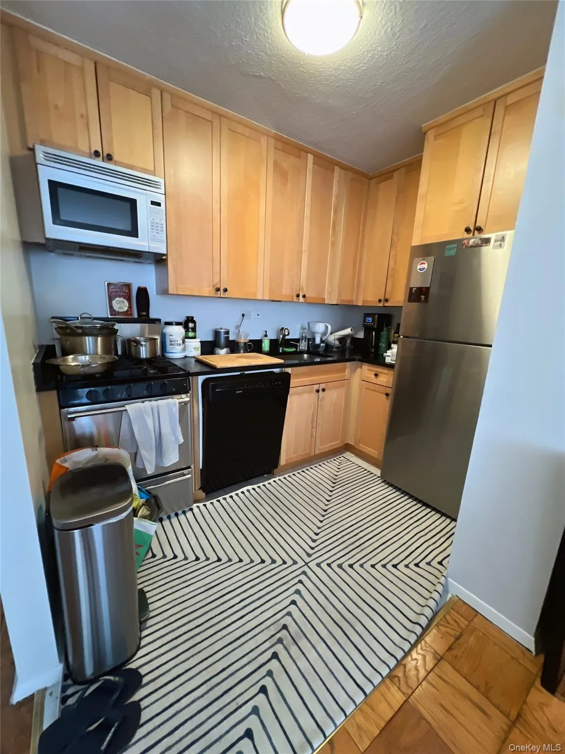 Kitchen featuring dark countertops, a textured ceiling, stainless steel appliances, and light brown cabinetry Kitchen featuring dark countertops, a textured ceiling, stainless steel appliances, and light brown cabinetry