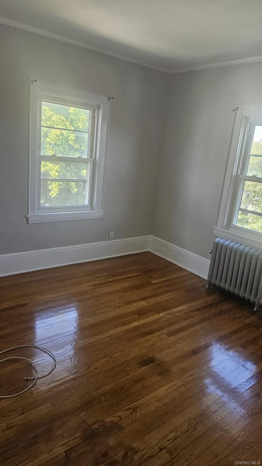 Empty room featuring radiator heating unit, crown molding, dark wood-style flooring, and healthy amount of natural light Empty room featuring radiator heating unit, crown molding, dark wood-style flooring, and healthy amount of natural light