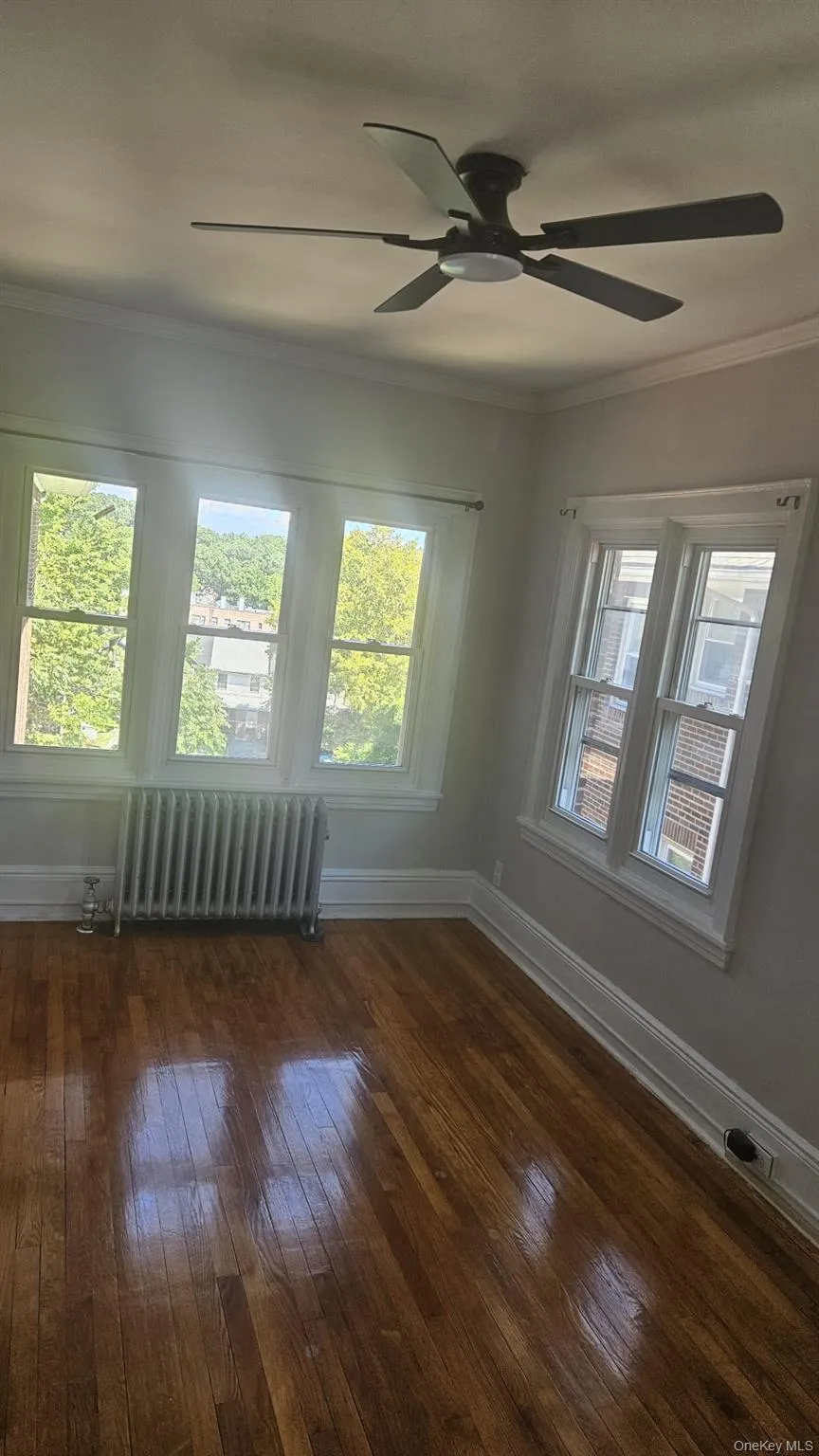 Unfurnished room featuring crown molding, dark wood finished floors, radiator heating unit, and ceiling fan Unfurnished room featuring crown molding, dark wood finished floors, radiator heating unit, and ceiling fan
