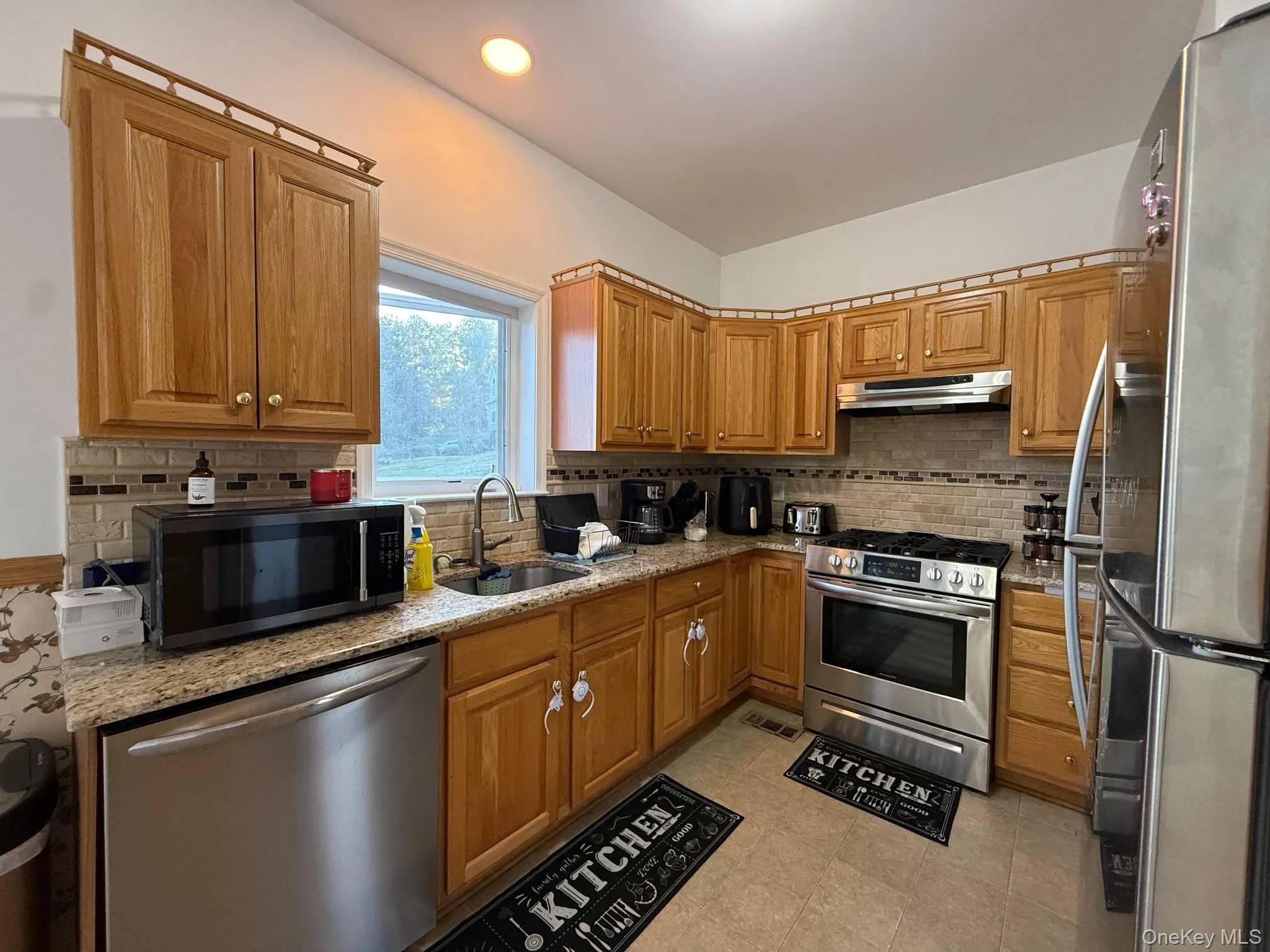 Kitchen featuring appliances with stainless steel finishes, brown cabinetry, tasteful backsplash, light stone countertops, and under cabinet range hood Kitchen featuring appliances with stainless steel finishes, brown cabinetry, tasteful backsplash, light stone countertops, and under cabinet range hood
