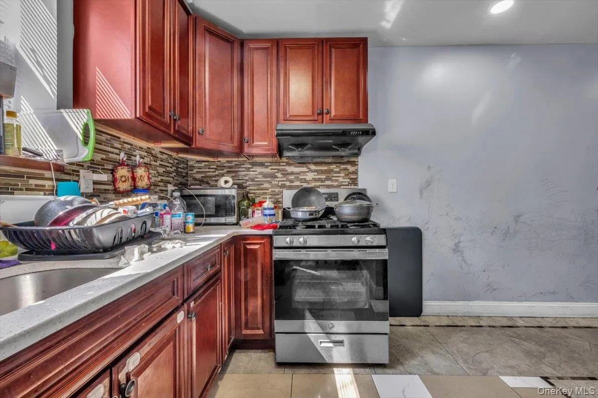 Kitchen featuring appliances with stainless steel finishes, tasteful backsplash, reddish brown cabinets, light stone countertops, and light tile patterned floors Kitchen featuring appliances with stainless steel finishes, tasteful backsplash, reddish brown cabinets, light stone countertops, and light tile patterned floors