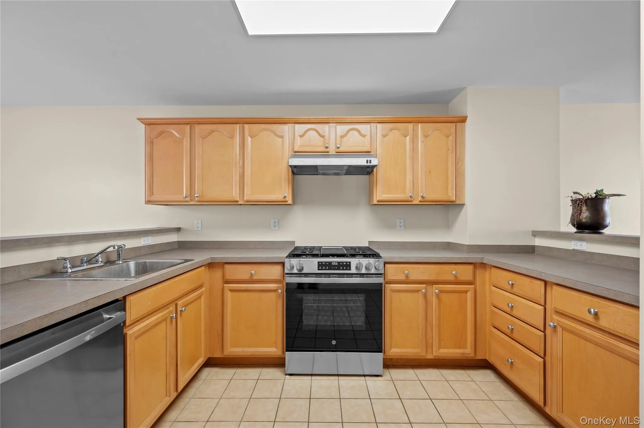 Kitchen with stainless steel appliances, light tile patterned floors, under cabinet range hood, light brown cabinets, and a skylight Kitchen with stainless steel appliances, light tile patterned floors, under cabinet range hood, light brown cabinets, and a skylight